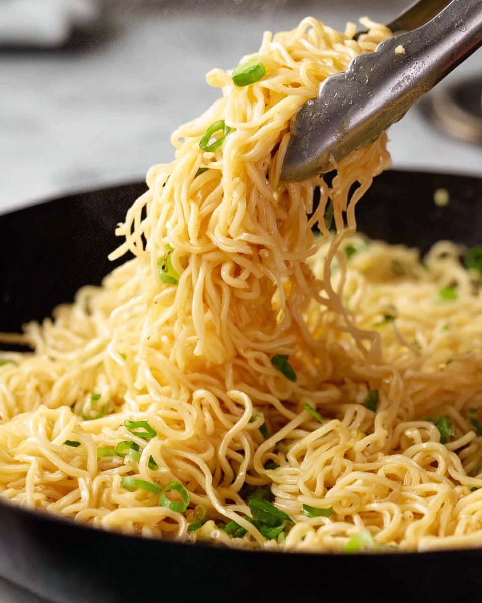 A close-up view shows a bundle of cooked yellow noodles with a slight shine, held by silver metal tongs. Small green pieces, likely chopped scallions, are scattered throughout the noodles. The noodles appear soft and tangled, with thin, wavy strands. The background features a black pan with more noodles and green bits visible, set on a white marbled surface. The photo captures the texture and slight gloss of the noodles clearly, showing steam rising slightly, indicating warmth. photo taken with an iphone --ar 4:5 --v 7