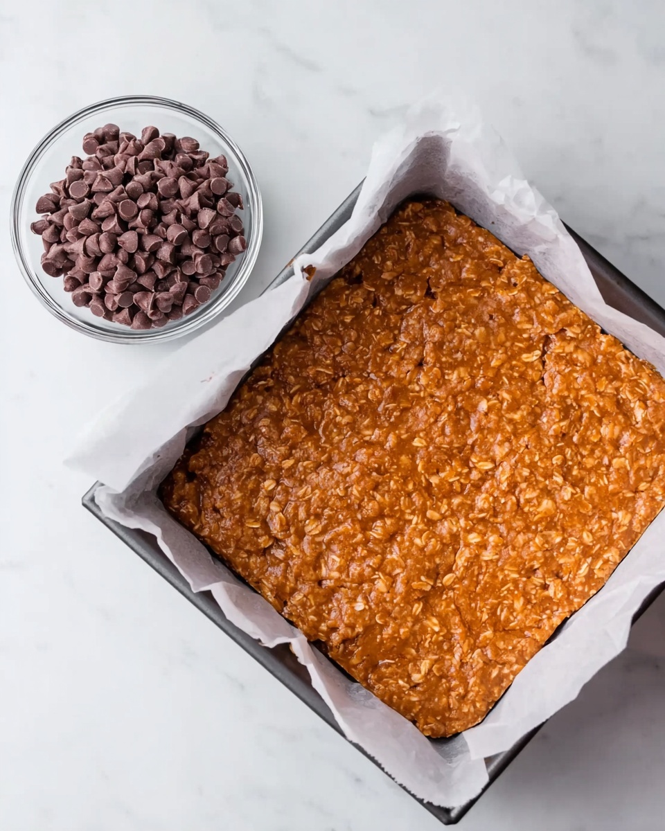 A square baking pan lined with white parchment paper holds a thick, textured, orange-brown mixture spread evenly to the edges. Next to the pan is a small clear bowl filled with smooth, dark brown chocolate chips. Both are placed on a white marbled surface. The mixture’s surface shows a rough, slightly bubbly texture that hints at oats or similar ingredients. Photo taken with an iphone --ar 4:5 --v 7