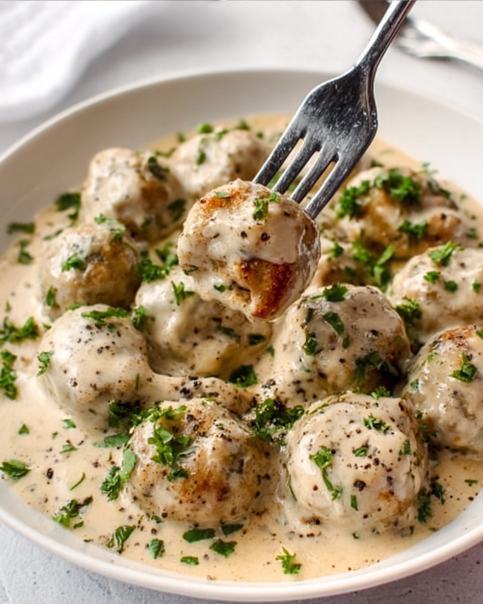 A white round plate filled with small round meatballs covered in a creamy beige sauce, sprinkled with fresh green chopped herbs and black pepper. A silver fork is lifting one meatball, showing its soft inside texture. The surface is a white marbled texture. photo taken with an iphone --ar 4:5 --v 7