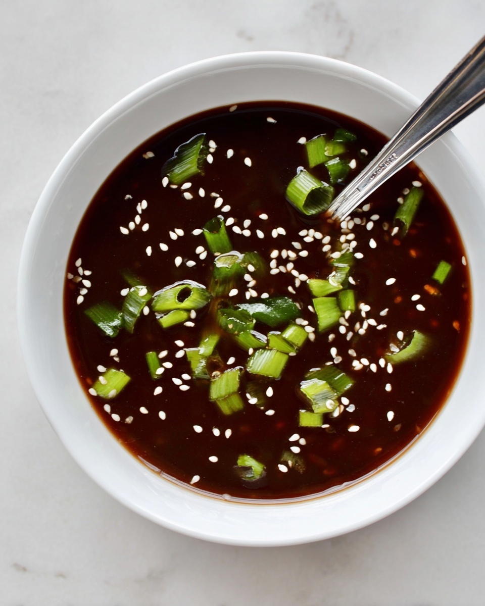 A white bowl filled with a dark brown sauce that looks smooth and thick, topped with small white sesame seeds evenly spread on the surface. Bright green chopped scallions are scattered on top, adding freshness and color contrast. A silver spoon is placed inside the bowl, partially immersed in the sauce. The bowl is set on a white marbled texture background. Photo taken with an iphone --ar 4:5 --v 7