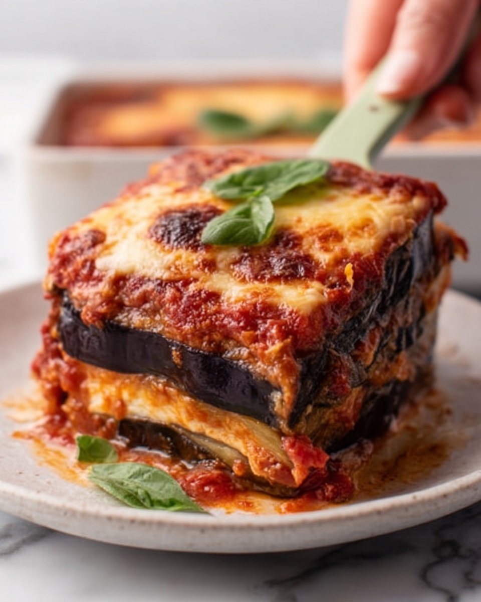 A white plate holds a thick slice of baked layered dish showing multiple layers; the bottom and middle layers are dark purple, smooth slices of eggplant, topped with a rich red tomato sauce layer covered by a golden-brown melted cheese layer with a slightly crispy top. Small green basil leaves decorate the top surface. A woman's hand is using a pale green spatula to lift the slice from a white marbled surface. Photo taken with an iphone --ar 4:5 --v 7