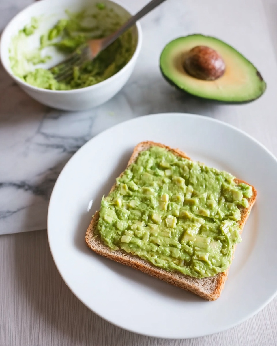 A white plate with one slice of toast covered evenly with a thick layer of light green mashed avocado, showing some small chunks for texture. Behind the plate, on a white marbled surface, there is a white bowl with some leftover mashed avocado inside and a fork resting in it. Next to the bowl is one half of an avocado with the seed still inside, showing a rich green color inside and a dark outer skin. Photo taken with an iphone --ar 4:5 --v 7