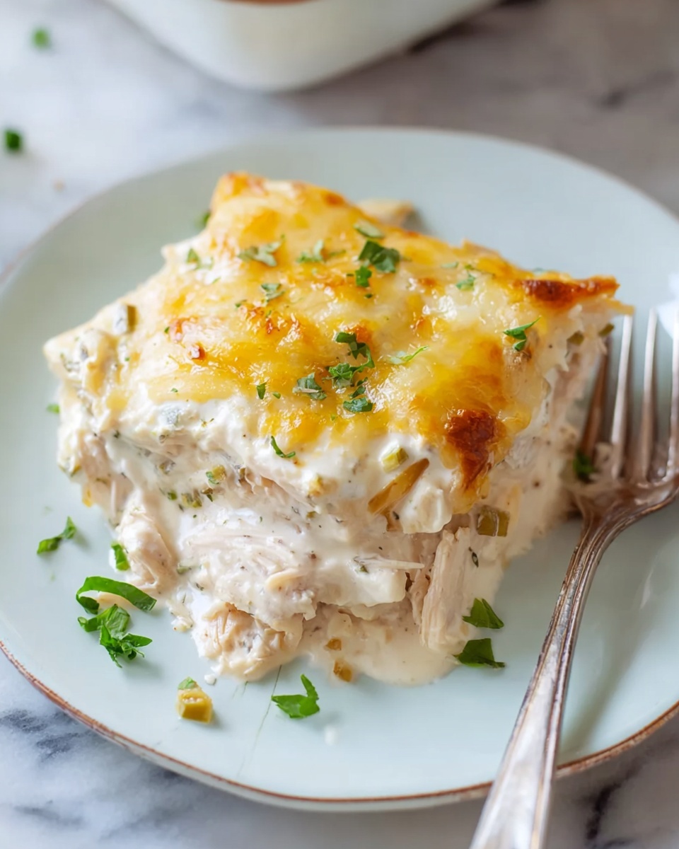 A single piece of layered creamy chicken casserole sits on a white plate with a light blue rim. The dish has three main layers: a bottom layer of shredded light beige chicken, a middle thick layer of creamy white sauce with small green pieces, and a top layer of soft melted pale yellow cheese with some golden brown spots. Small green herb pieces are sprinkled on top and around the plate. A silver fork rests on the right side of the plate. The background surface is white marble. Photo taken with an iphone --ar 4:5 --v 7