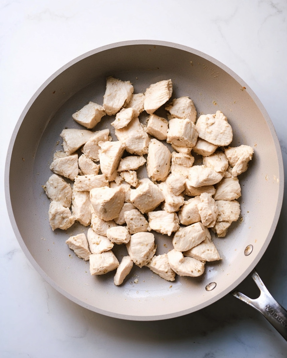 A top view of a light gray frying pan with many small, irregular pieces of cooked white chicken placed inside. The pieces are spread mostly evenly around the pan with no overlapping. The pan is on a white marbled surface, and the pan handle is partially visible on the right side. The chicken pieces have a slightly rough texture and a pale beige color. Photo taken with an iphone --ar 4:5 --v 7