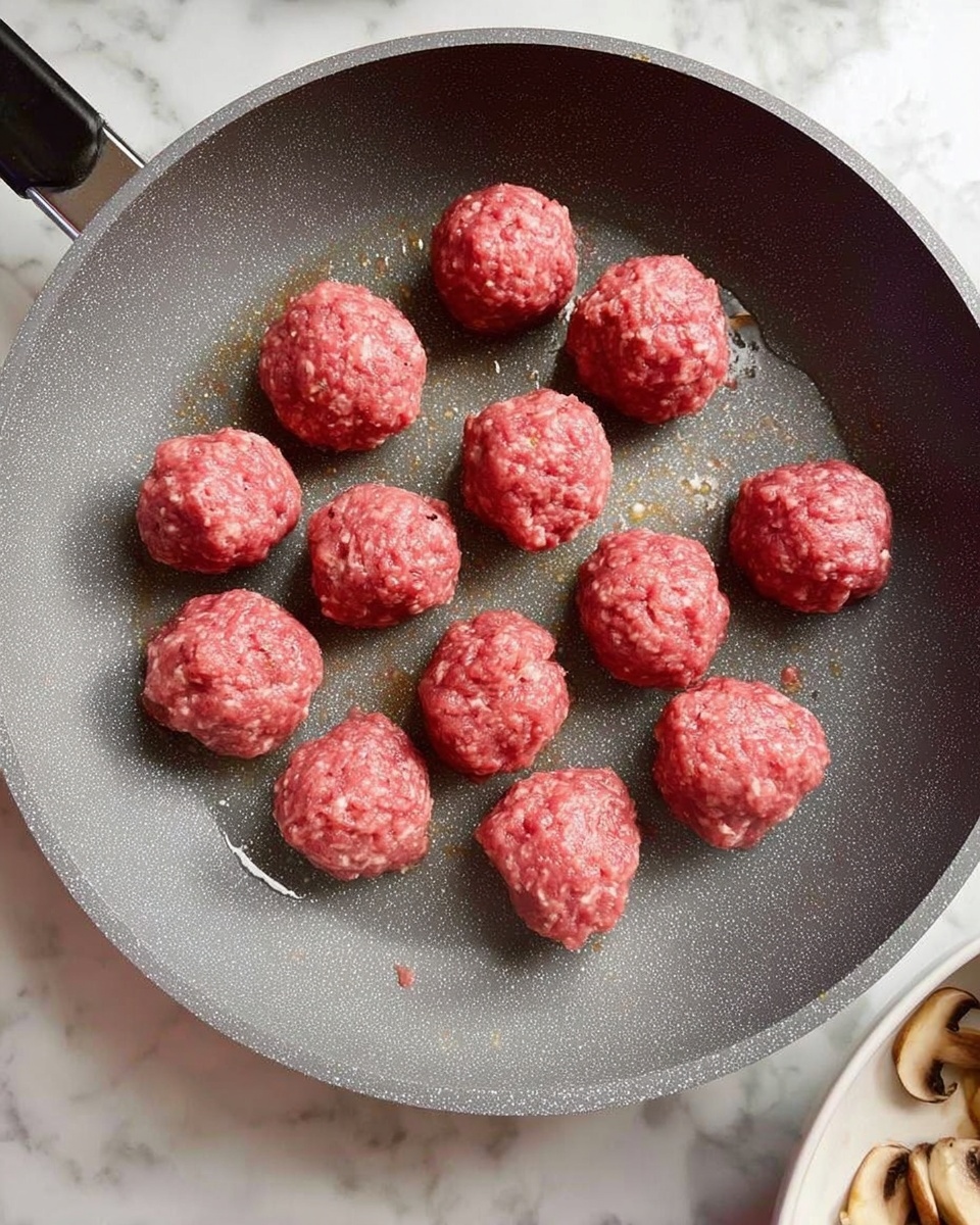 The image shows a gray non-stick frying pan on a white marbled surface. Inside the pan, there are eleven small, round raw meatballs, evenly spaced apart. The meatballs have a pinkish-red color, with a slightly rough texture made from ground meat. Some oil is visible around the meatballs, giving a shiny effect in parts of the pan. The edge of a white bowl filled with mushrooms and some other ingredients is seen in the bottom right corner. Photo taken with an iphone --ar 4:5 --v 7