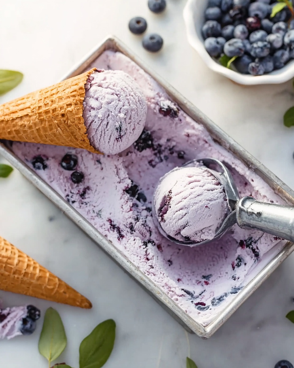 The image shows a metal rectangular container filled with light purple ice cream mixed with small dark purple bits, likely blueberries. A metal ice cream scoop is inside the container, holding a rounded scoop of the ice cream. Next to it, a waffle cone lies on the edge of the container, filled with a similarly colored scoop of the ice cream. In the background, there is a white bowl with blueberries and some loose blueberries and green leaves on a white marbled surface. Photo taken with an iphone --ar 4:5 --v 7