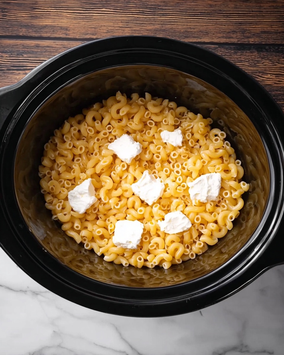 A black slow cooker filled with cooked golden macaroni pasta that has a soft and slightly shiny texture. Scattered on top are five uneven white cream cheese dollops, contrasting with the yellow pasta. The cooker sits on a white marbled surface, showing a smooth, rustic wooden background but adjusted here as white marble for the visual prompt. photo taken with an iphone --ar 4:5 --v 7