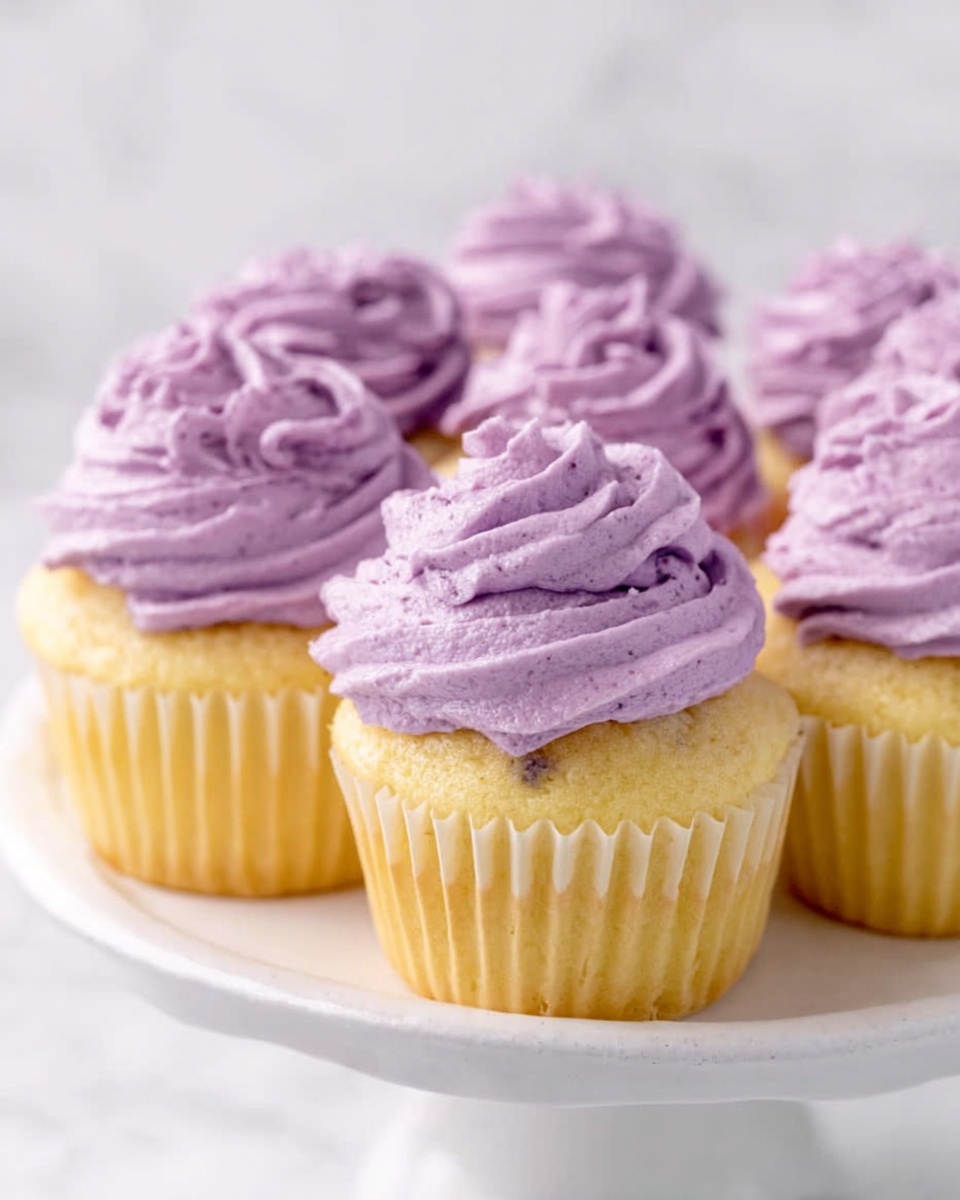 The image shows a group of yellow cupcakes arranged closely together on a white cake stand with delicate paper liners. Each cupcake has one thick layer of smooth, creamy, purple frosting swirled on top with a slightly textured appearance, giving a soft and fluffy look. The background and surface beneath the cake stand feature a white marbled texture, adding a clean and bright atmosphere to the scene. The photo looks sharp and clear, taken with an iphone --ar 4:5 --v 7