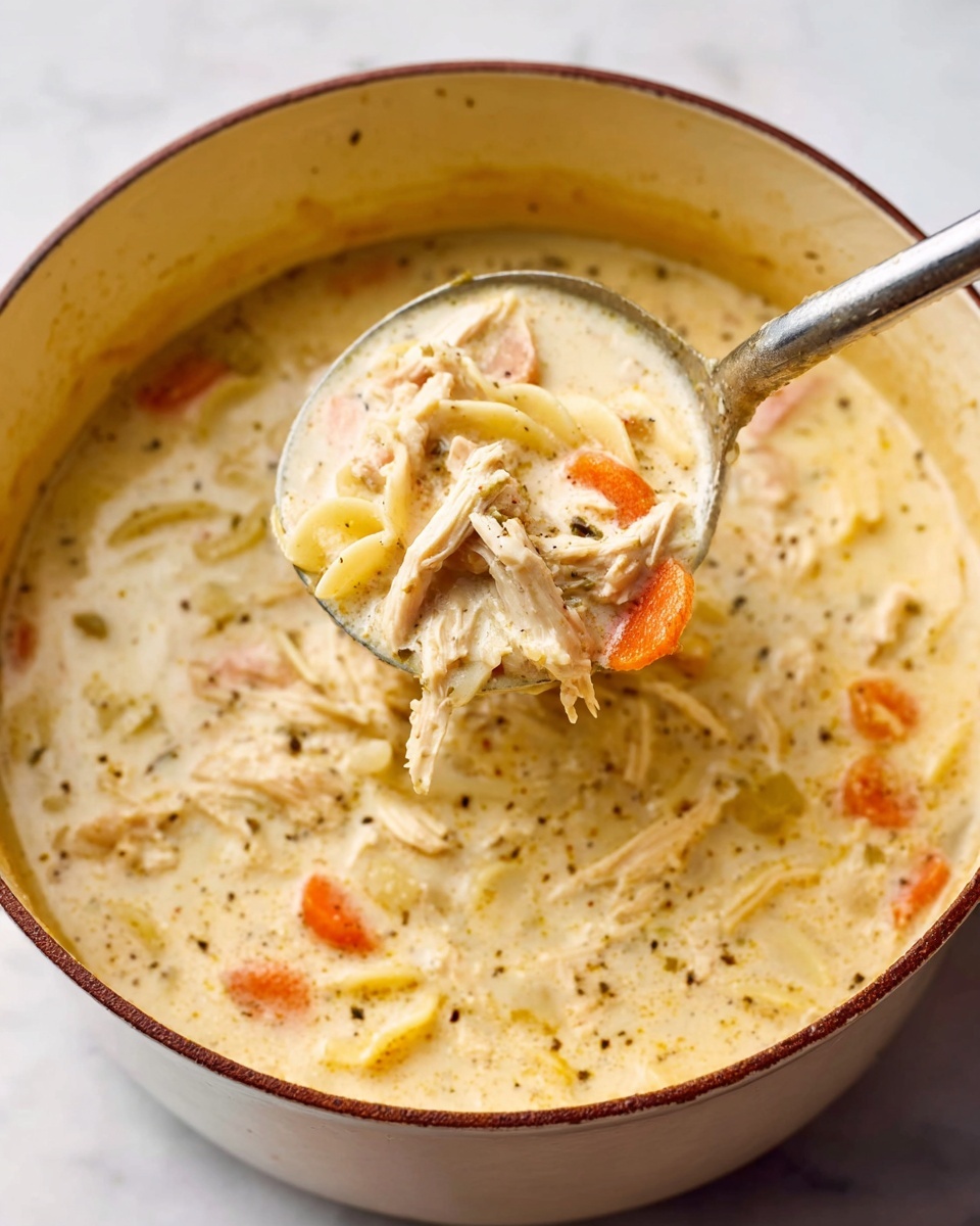 The image shows a close-up of creamy chicken soup in a white pot with a brown rim. The soup has a thick, smooth texture with visible shredded white chicken layers, soft small orange carrot slices, and light yellow pasta bits floating evenly throughout. There are small black pepper flakes sprinkled on the surface. A metal ladle scoops the soup, showing more layers of the creamy broth and ingredients dripping slowly back into the pot. The pot sits on a white marbled surface, and the overall color palette is warm and comforting with light cream, orange, and brown tones. Photo taken with an iphone --ar 4:5 --v 7