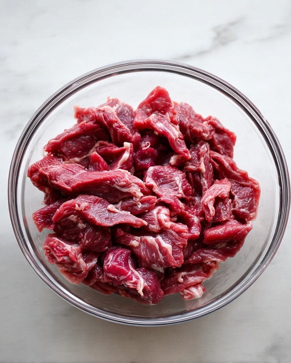A clear glass bowl sits on a white marbled surface, filled with thin, uneven strips of raw red meat. The meat has visible fibrous texture and some white fat streaks mixed throughout, creating a contrast of deep red and pale tones. The bowl's transparency shows the meat from the sides and below, with natural light highlighting the wet texture of the meat strands. photo taken with an iphone --ar 4:5 --v 7
