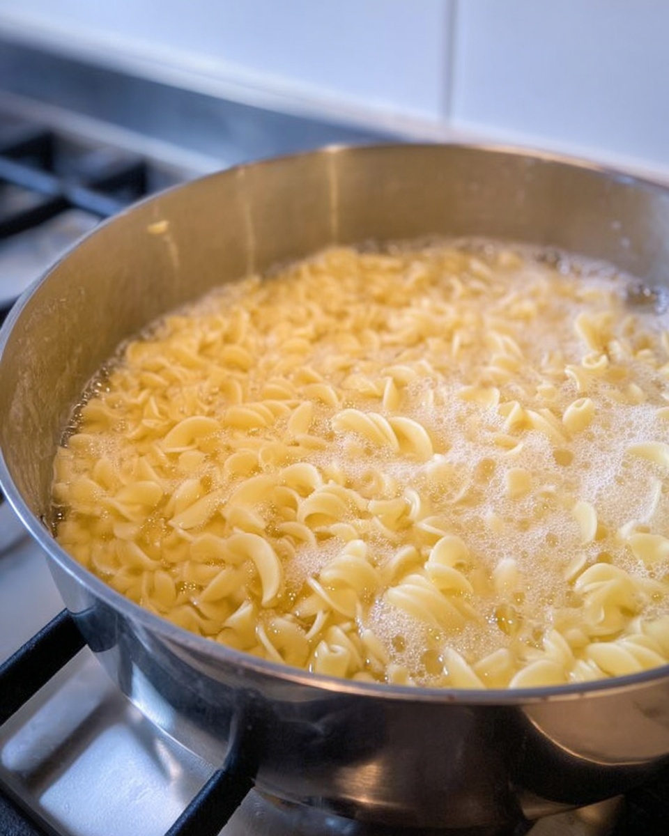 A close-up view of a shiny silver pot filled with pale yellow curly pasta boiling in clear bubbling water with white foam on the surface. The pot sits on a gas stove with silver grates, and the background shows parts of the white kitchen wall. The texture of the pasta is soft and wavy as it cooks in the water. photo taken with an iphone --ar 4:5 --v 7