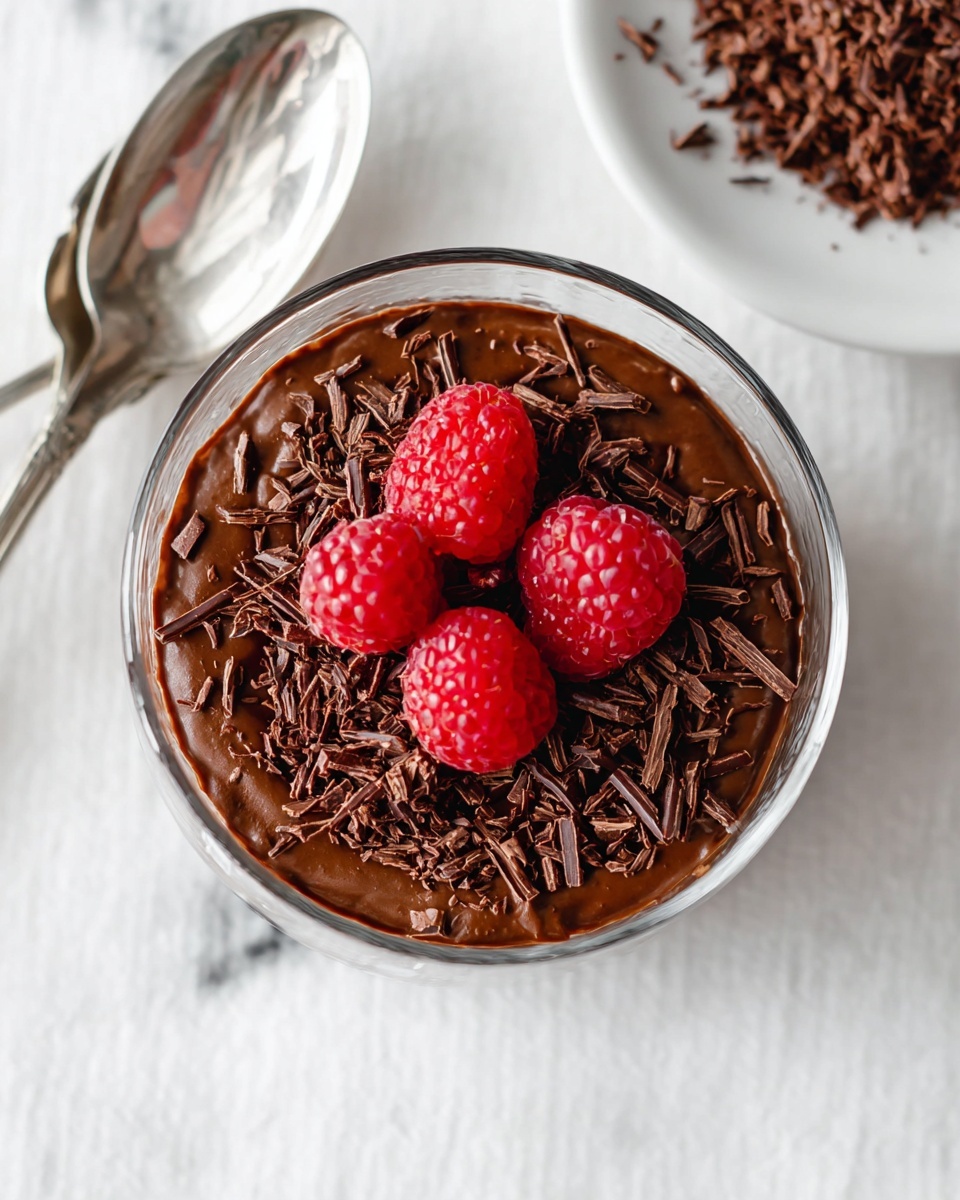 A clear glass bowl filled with a thick, dark brown chocolate pudding layer, topped with shaved dark chocolate pieces scattered on top, and four bright red frozen raspberries arranged in the center. The bowl is placed on a white marbled surface, with a silver spoon to the left. At the top edge of the frame, a white plate with additional dark chocolate shavings is partially visible. Photo taken with an iphone --ar 4:5 --v 7