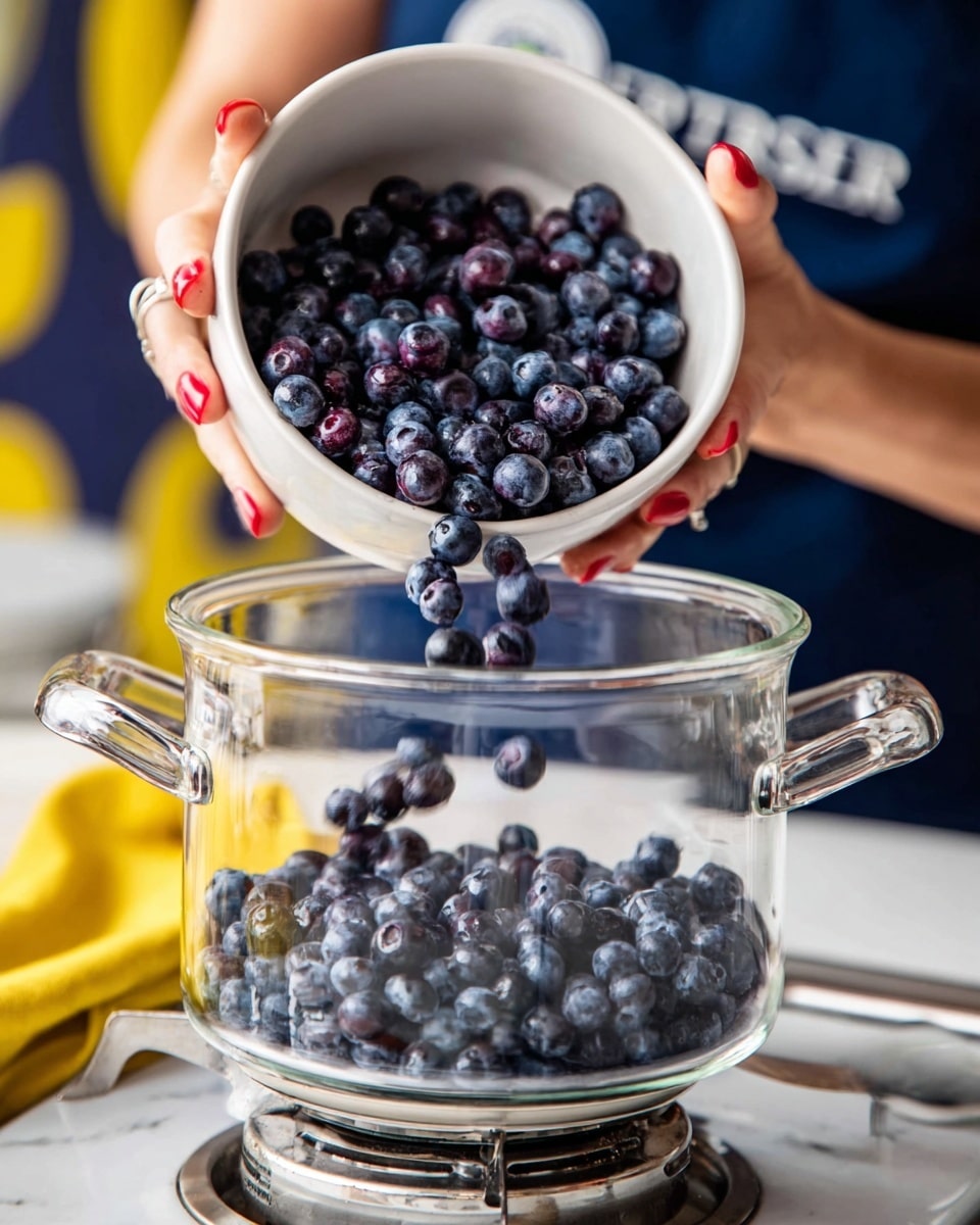 A clear glass pot with two handles is placed on a silver stove burner, on a white marbled surface. A white bowl overflowing with fresh, shiny dark blue blueberries is held by a woman's hand with red nail polish, as blueberries pour from the bowl into the pot. The blueberries inside the bowl and those dropping into the pot show a mix of deep blue and slight purple shades with a smooth texture. The background has blurred elements of a person wearing navy blue clothes and a yellow cloth. Photo taken with an iphone --ar 4:5 --v 7