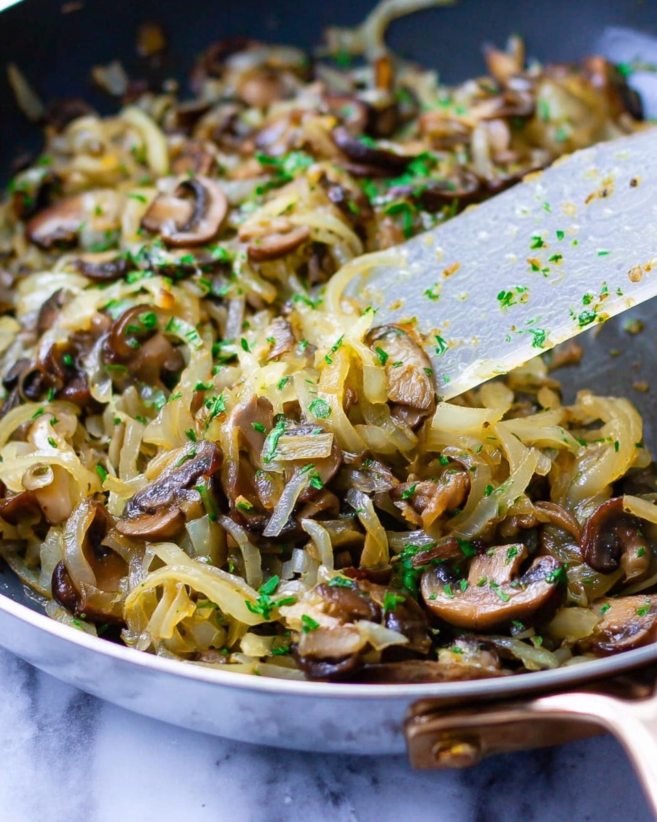 The image shows a close-up of a pan filled with cooked onions and sliced mushrooms mixed together. The onions are soft and light golden with a slightly translucent look, intertwined with brown and darker brown mushrooms that have a tender texture. The dish is sprinkled with chopped green herbs, adding a fresh color contrast. A translucent spatula with bits of the mixture clinging to it is visible in the pan, partially stirring the ingredients. The pan handle is slightly blurry in the foreground, resting on a white marbled surface. Photo taken with an iphone --ar 4:5 --v 7