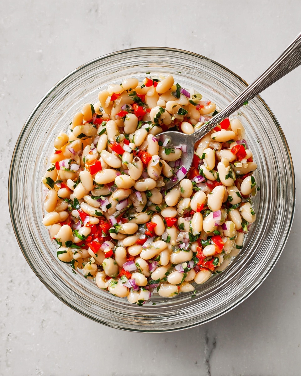 A clear glass bowl sits on a white marbled surface, filled with a mixed bean salad made mostly of white beans. The salad has small chopped pieces of red bell pepper, green herbs, and bits of light purple onion scattered throughout, adding color and texture. A silver spoon rests inside the bowl, partially buried in the beans and salad mix. The bowl’s transparent sides show the ingredients clearly with a slight shine from a dressing coating the beans. Photo taken with an iphone --ar 4:5 --v 7