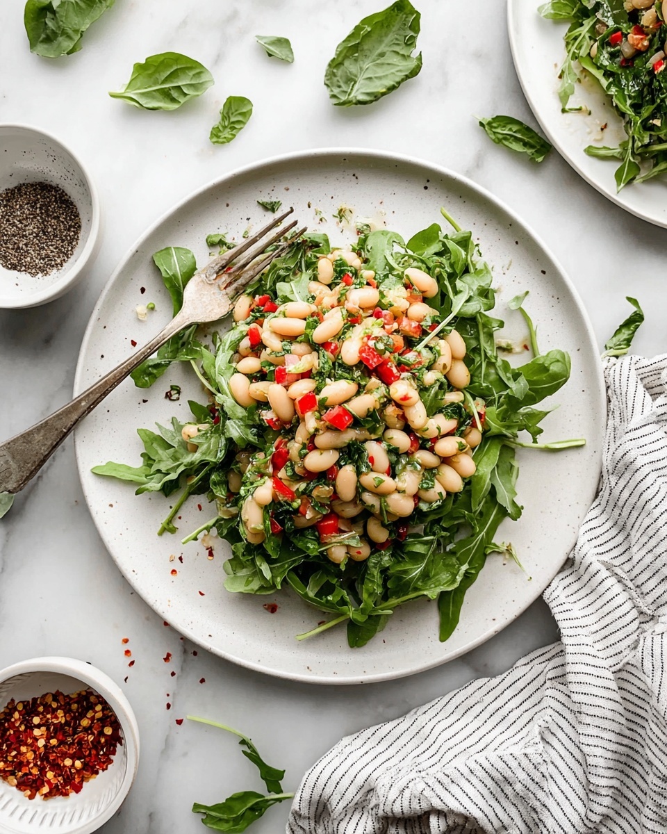 The image shows a white plate on a white marbled surface with a salad. The salad has two main layers: the bottom layer is fresh green arugula leaves spread unevenly on the plate. On top, there is a mix of white beans, small pieces of red bell pepper, chopped herbs, and finely diced onions, all lightly dressed with a shiny vinaigrette. A silver fork with some salad on it rests on the left side of the plate. Around the plate, there are some green leaves scattered, a striped cloth napkin to the bottom left, and two small white bowls, one with red chili flakes and the other with cracked black pepper. Photo taken with an iphone --ar 4:5 --v 7