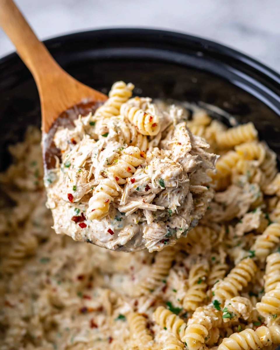 The image shows a close-up of a wooden spoon lifting a scoop of creamy pasta from a black pot. The pasta is rotini, light golden color, mixed with shredded chicken coated in a thick white sauce. There are green herbs and red spice flakes sprinkled throughout the dish, adding small pops of color. The sauce looks rich and smooth, covering each pasta piece and chicken shred evenly. In the pot below, the pasta mixture fills the space with a comforting, hearty texture. The scene is set against a white marbled surface. photo taken with an iphone --ar 4:5 --v 7