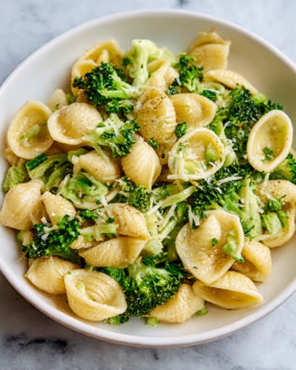 The image shows a white bowl filled with shell-shaped pasta mixed evenly with small green broccoli florets. The pasta pieces are light yellow with a smooth texture, and the broccoli adds a fresh green color that complements the dish. The pasta and broccoli are coated with a light, glossy sauce that gives the dish a moist look. The bowl is placed on a white marbled surface, and a woman's hand is reaching from the bottom edge towards the bowl. photo taken with an iphone --ar 4:5 --v 7