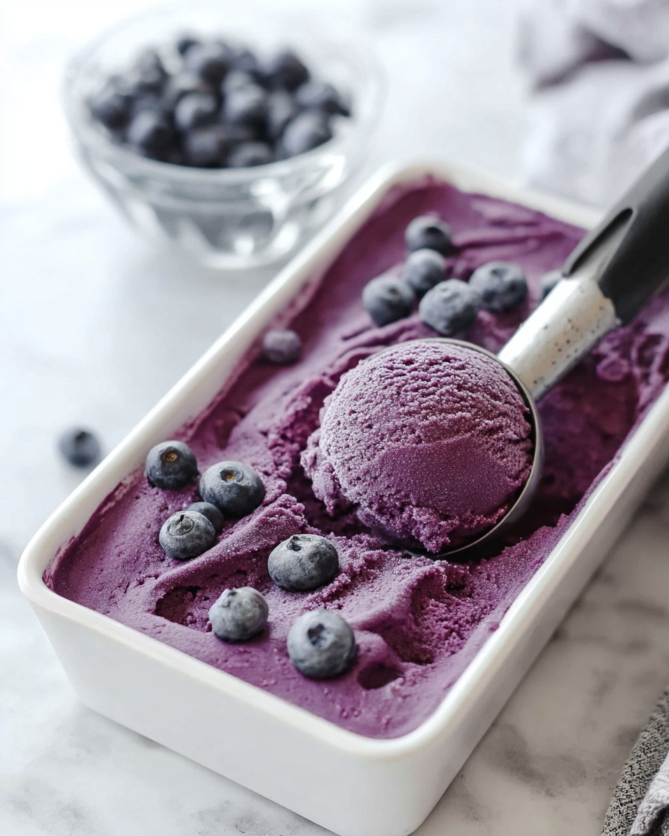 A white rectangular tray filled with smooth, deep purple blueberry ice cream with a creamy texture. The surface is decorated with whole frosted blueberries scattered evenly on top. A silver ice cream scoop with a black handle is lifting a round, perfectly shaped scoop of the ice cream from the tray. In the background, there is a small clear glass bowl filled with fresh blueberries on a white marbled surface. photo taken with an iphone --ar 4:5 --v 7