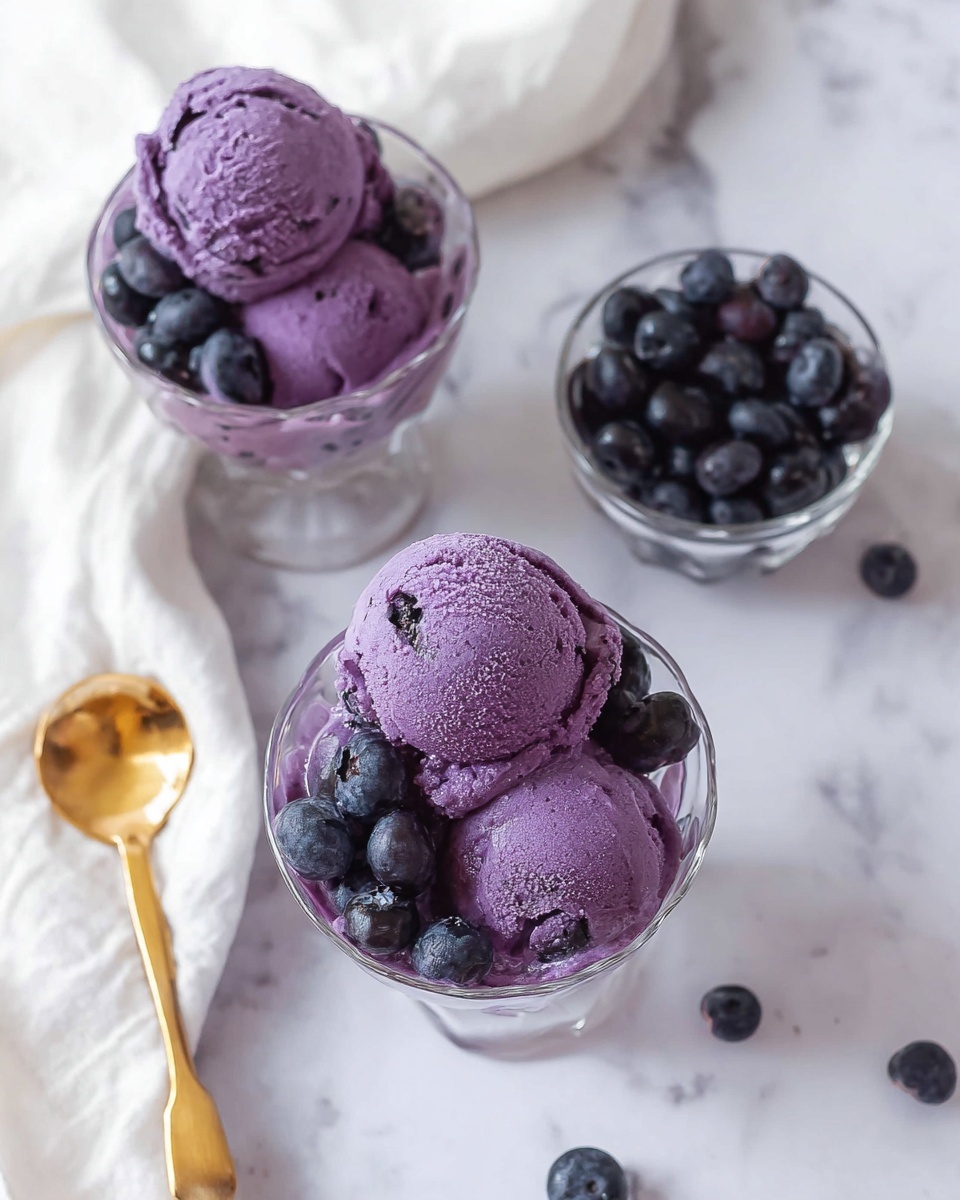 The image shows three glass bowls on a white marbled surface. Two bowls contain three scoops each of purple ice cream with a smooth, slightly textured surface, topped and surrounded by fresh dark blueberries. The third bowl is filled only with dark blueberries. A shiny gold spoon is visible near the bottom left, next to a white cloth. The overall colors contrast with the purple ice cream and dark berries against the white background. Photo taken with an iphone --ar 4:5 --v 7