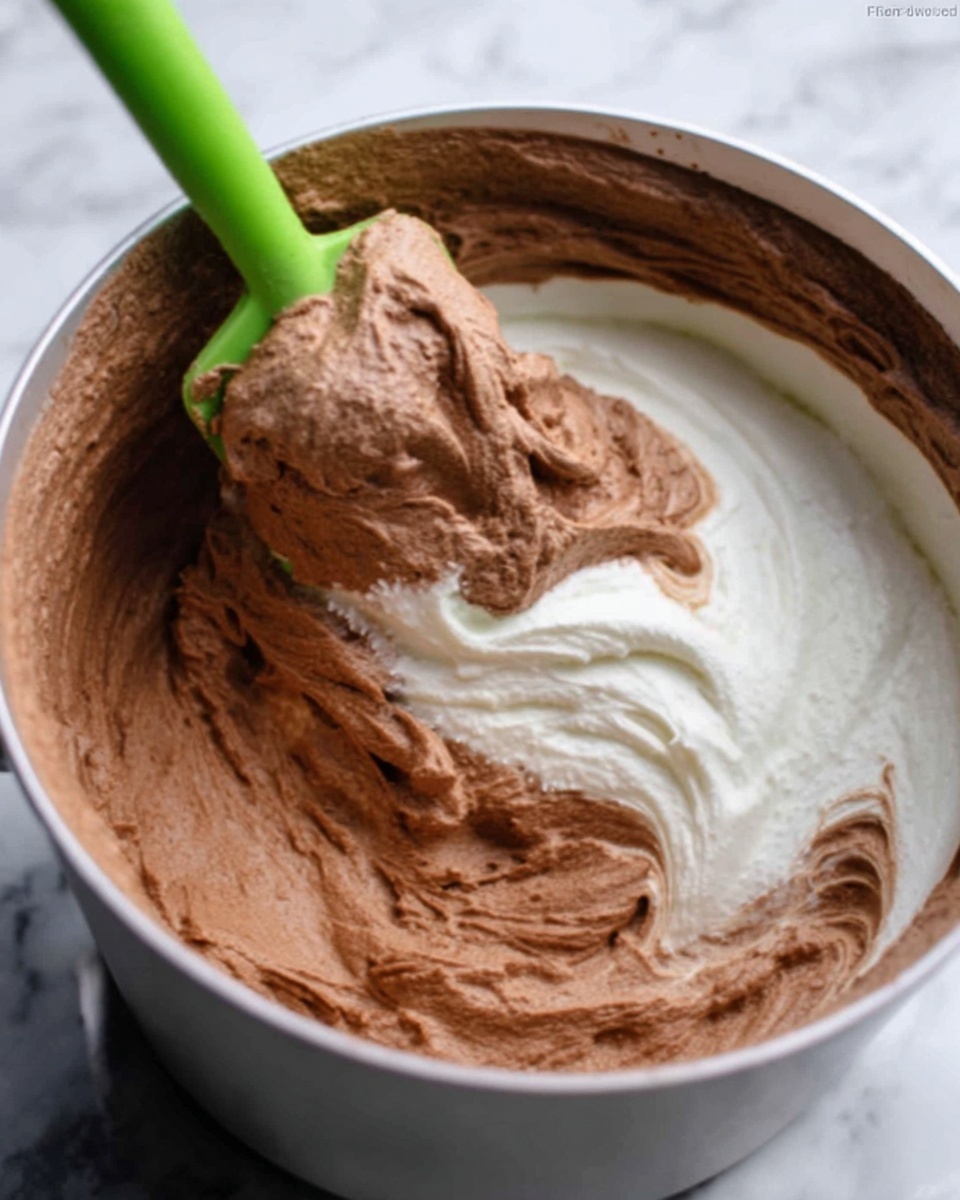 The image shows a close-up of a white pot with two layers of thick mixtures inside. The bottom layer is a smooth, white cream, while the top layer is a fluffy light brown chocolate mixture. A green spatula is lifting the brown mixture, mixing it gently with the white cream. The textures look soft and creamy, and the focus is on the mixing motion. The background is a white marbled texture. Photo taken with an iphone --ar 4:5 --v 7