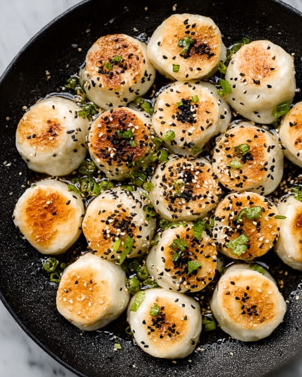 The image shows a black round pan filled with small, round dumplings that have golden brown tops, with the rest of their soft, white dough visible. The dumplings are arranged closely together, some sprinkled lightly with black sesame seeds and green chopped herbs, adding touches of black and green color on top. The pan sits on a white marbled surface with small droplets of oil shining around the dumplings. photo taken with an iphone --ar 4:5 --v 7