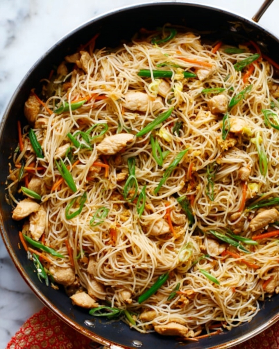 This image shows a close-up of a black pan filled with cooked thin noodles mixed with small pieces of light brown chicken. The noodles are a pale beige color and look soft and slightly oily. There are thin green slices of spring onions spread across the top, adding color and texture. Some thin orange strips of carrot are mixed in among the noodles and chicken, giving a subtle contrast. The pan is sitting on a white marbled surface. The photo is well-lit and looks natural. photo taken with an iphone --ar 4:5 --v 7