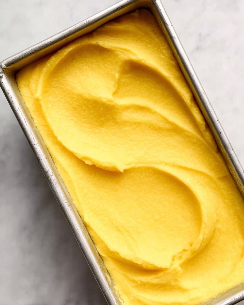 This is a close-up image of a rectangular silver metal tray filled with a smooth, bright yellow mixture that looks like soft mashed potatoes or polenta. The surface has gentle swirls and ridges made by a utensil, showing a creamy and thick texture. The tray is placed on a white marbled surface. photo taken with an iphone --ar 4:5 --v 7