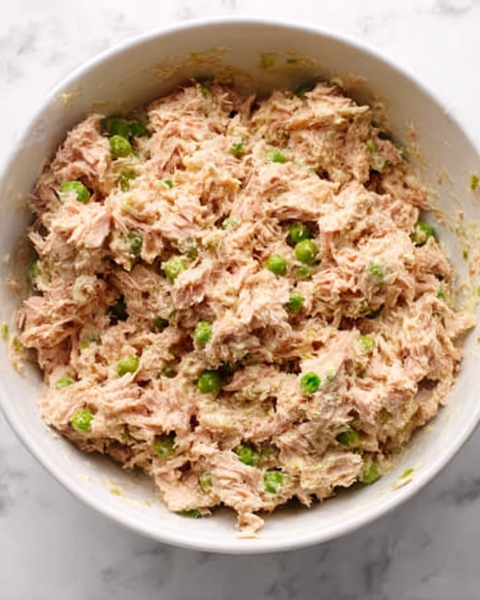 A close-up view of a white bowl filled with a creamy mixture of shredded light brown and pink tuna fish, mixed with small green peas and finely chopped onions. The texture looks soft with bits of vegetables scattered evenly throughout. The bowl is placed on a white marbled surface, making the colors of the tuna salad stand out clearly. Photo taken with an iphone --ar 4:5 --v 7