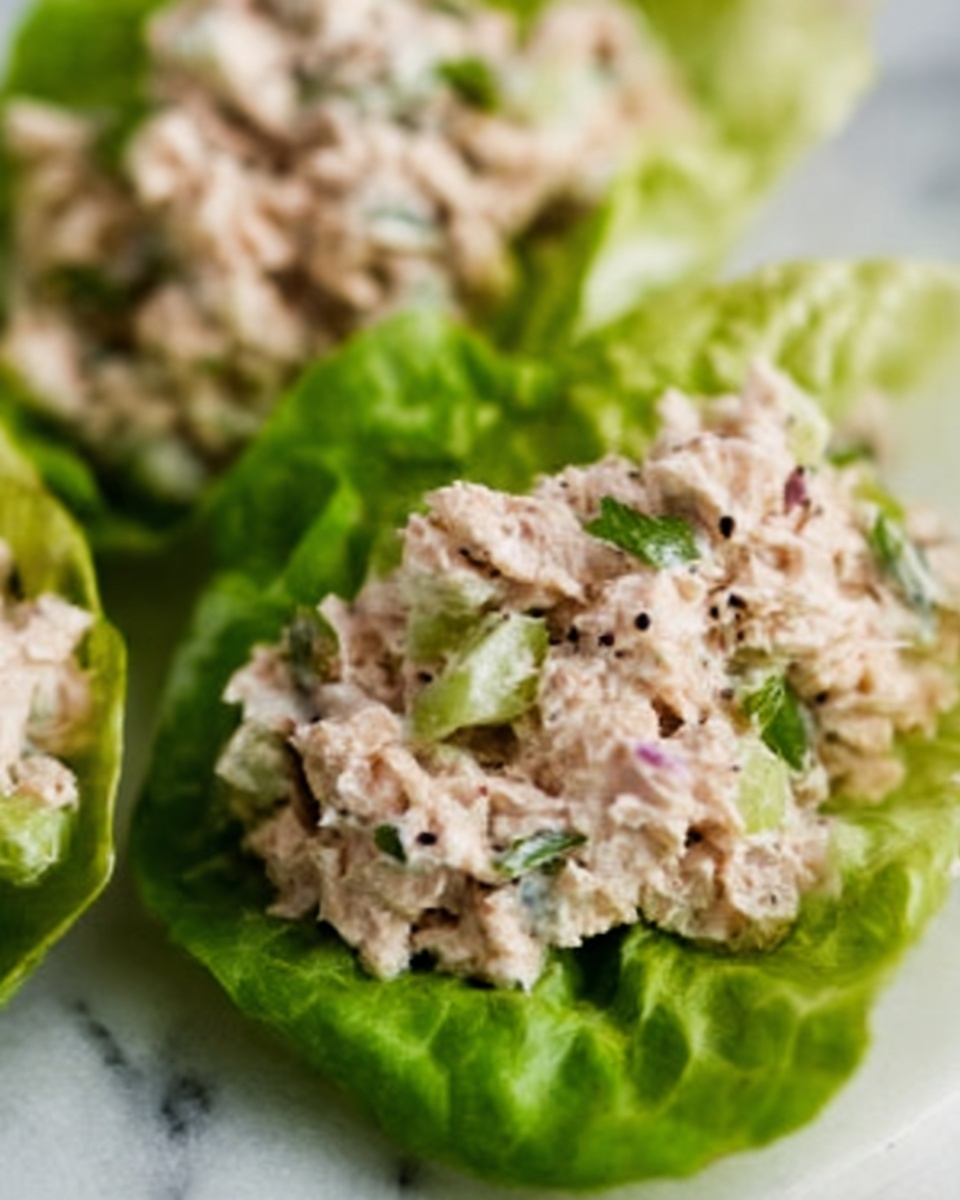 There are three green lettuce leaves on a white plate lined with parchment paper. Each lettuce leaf holds a mix of finely chopped white and pale pink meat, mixed with small pieces of purple onion and green herbs. The filling has a soft, slightly crumbly texture with black pepper sprinkled on top. In the blurred background, a small white bowl with a coarse reddish-brown sauce is visible. The setting has a clean white marbled surface. photo taken with an iphone --ar 4:5 --v 7