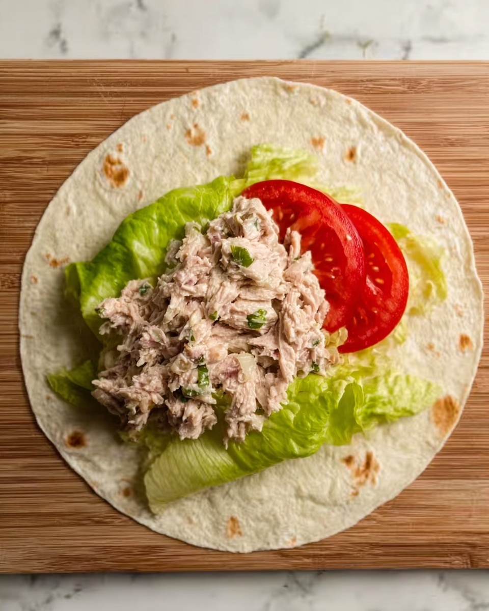 A large round tortilla lies flat on a wooden board with a white marbled texture beneath it. On the lower half of the tortilla, there is a base layer of green leafy lettuce, topped with a slice of fresh red tomato. Above the tomato and lettuce is a thick layer of shredded, light brown tuna mixed with small bits of green herbs, spread unevenly in the center. The tortilla is soft with small brown spots scattered across the surface. The scene is lit naturally, showing the textures and colors clearly. Photo taken with an iphone --ar 4:5 --v 7