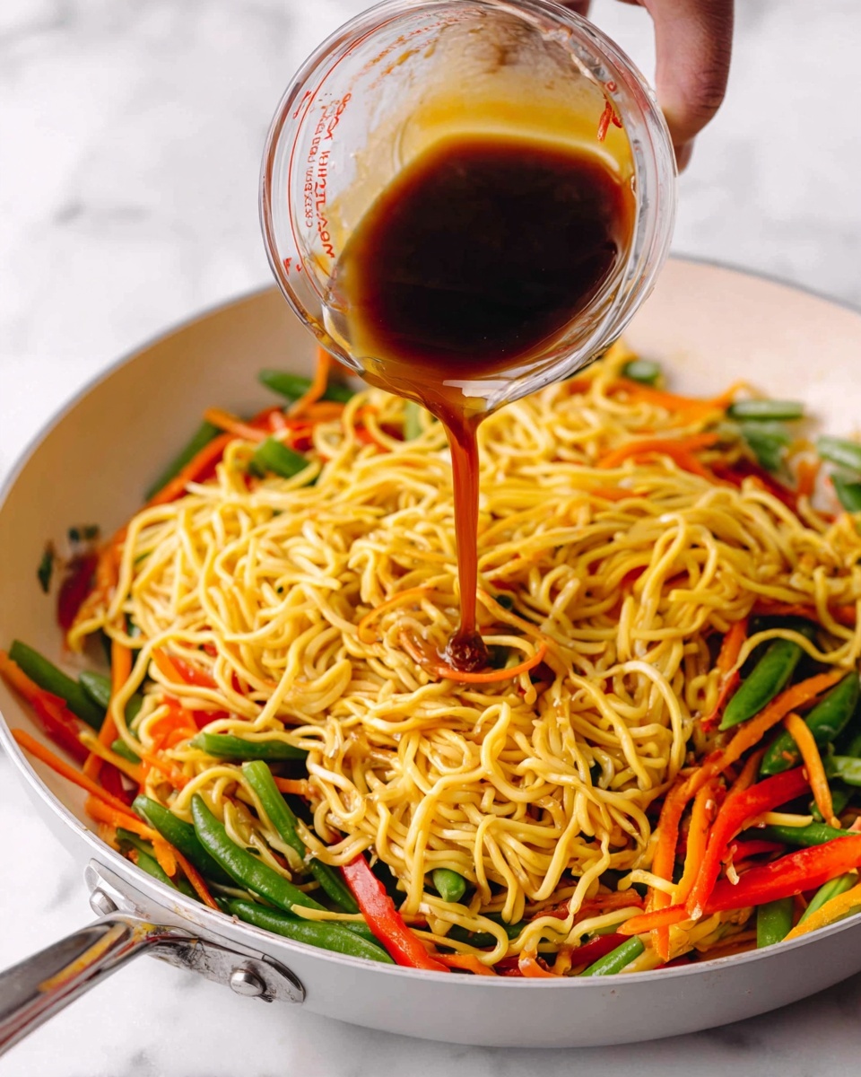 This image shows a white pan filled with cooked yellow noodles mixed with green snap peas and strips of red and orange vegetables like bell peppers and carrots. A woman's hand is pouring dark brown sauce from a clear measuring cup into the pan, with steam rising slightly. The pan is placed on a white marbled surface, and the colorful vegetables add bright contrasts to the scene. photo taken with an iphone --ar 4:5 --v 7