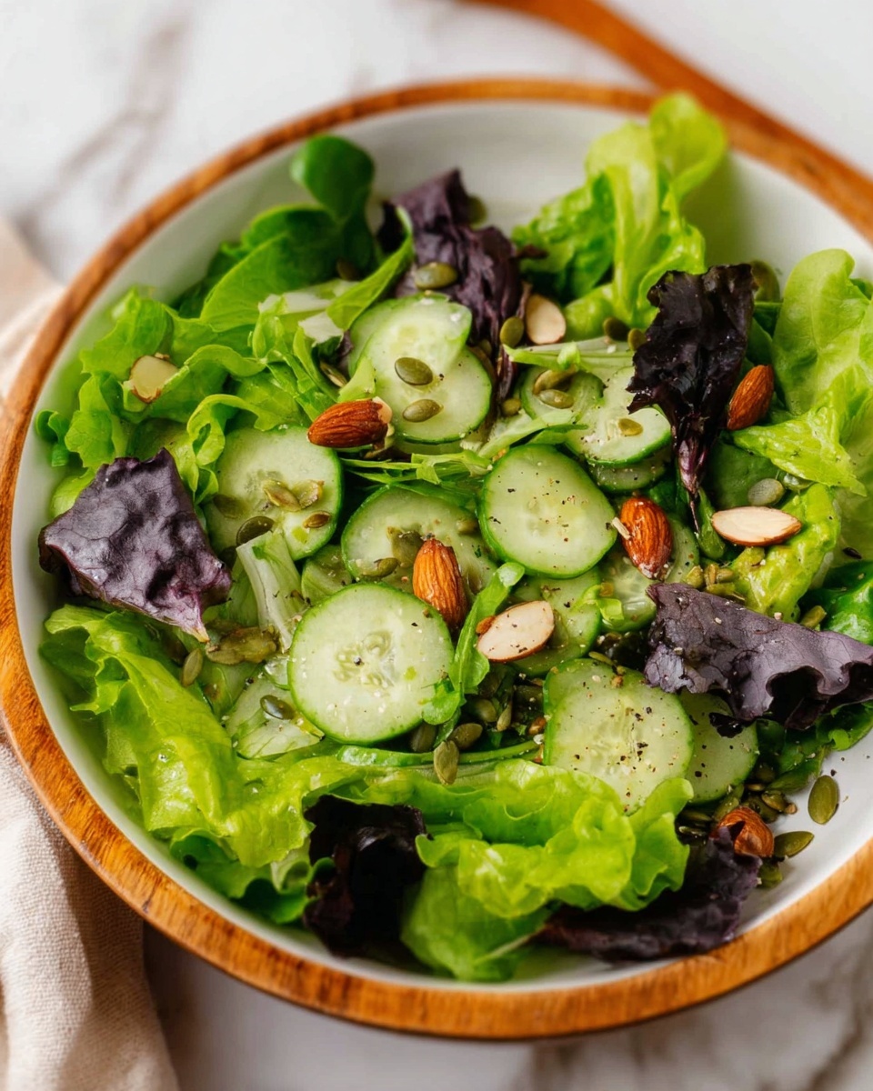 A fresh salad is layered in a white bowl with a wooden rim, placed on a white marbled surface. The bottom layer shows bright green curly lettuce leaves, some dark purple leaves mixed in. On top, there are thin, round slices of cucumber with green skins, scattered almonds, and small green pumpkin seeds. The salad has a light dressing with tiny specks of black pepper visible. The colors vary from light green to dark green, with the almonds adding a brown touch throughout. Photo taken with an iphone --ar 4:5 --v 7