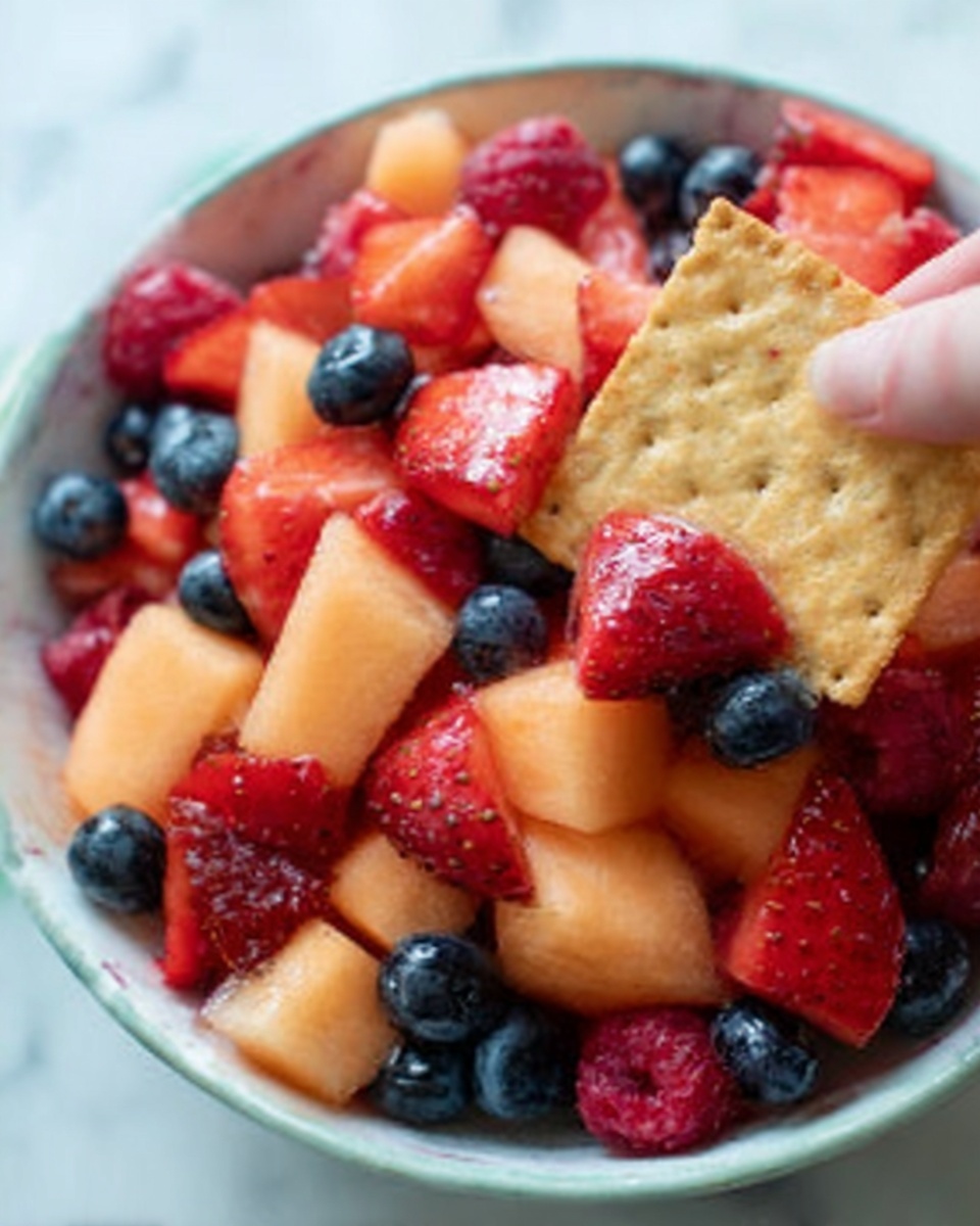 A close-up view of a white bowl on a white marbled surface filled with a colorful fruit salad. The salad has three visible layers: the bottom layer shows pale orange cantaloupe cubes with a smooth texture, the middle layer has bright red strawberries cut into small pieces, and the top layer features a mix of whole blueberries and whole red raspberries, adding dark blue and bright red colors. A woman's hand is holding a square cracker dipped into the fruit salad from the right side of the frame, with some fruit resting on the cracker's surface. Photo taken with an iphone --ar 4:5 --v 7
