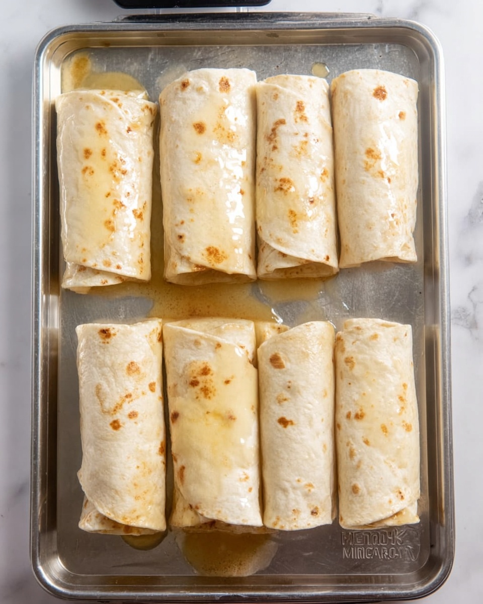 The image shows six rolled tortillas placed neatly on a flat metal baking tray. Each tortilla roll has a light beige color with small brown spots, covered with a shiny layer of what looks like sauce or melted butter, creating glistening highlights. The rolls are arranged in two rows of three, with some sauce spilled onto the tray around them. The surface beneath the tray is a white marbled texture with soft, subtle vein patterns. photo taken with an iphone --ar 4:5 --v 7
