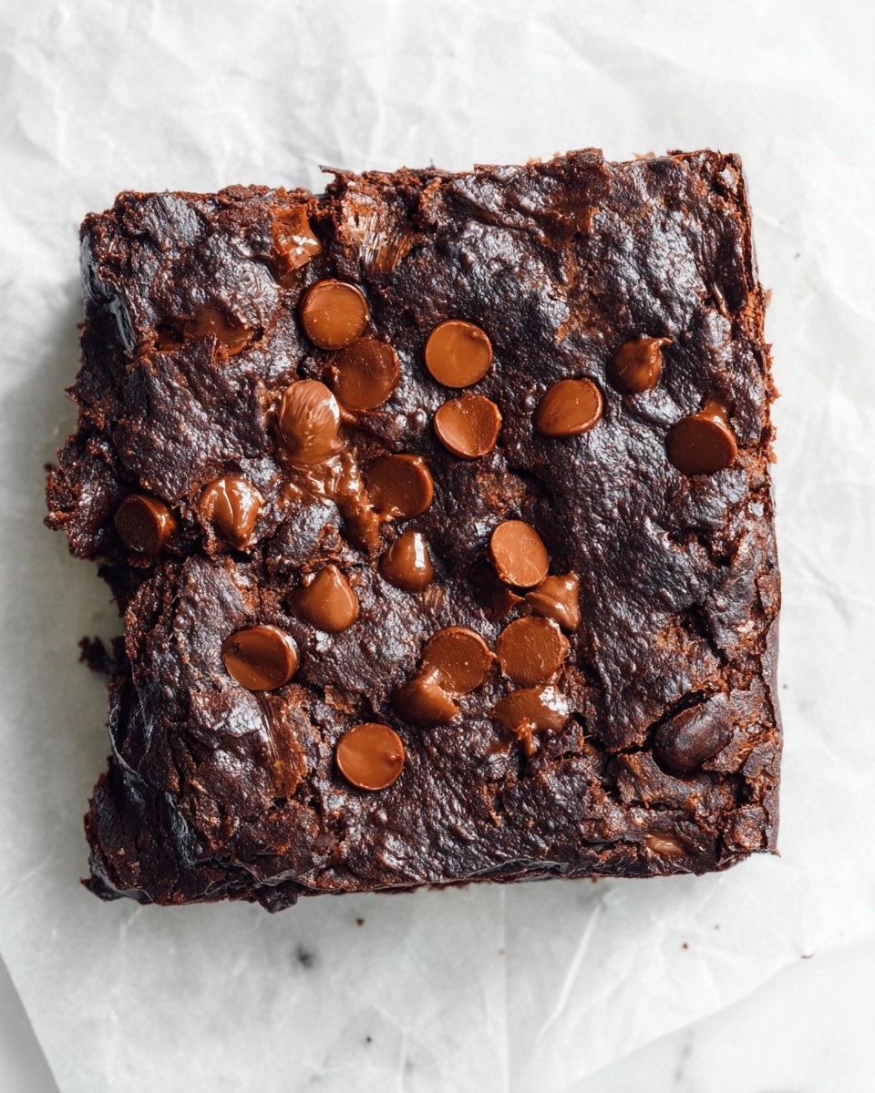 A square chocolate brownie with a rough, cracked dark brown surface and shiny, melted chocolate chips scattered unevenly on top. The brownie has a dense texture with small cracks revealing a rich, moist inside. It is placed on a white marbled background with a slightly crumpled white paper underneath. photo taken with an iphone --ar 4:5 --v 7