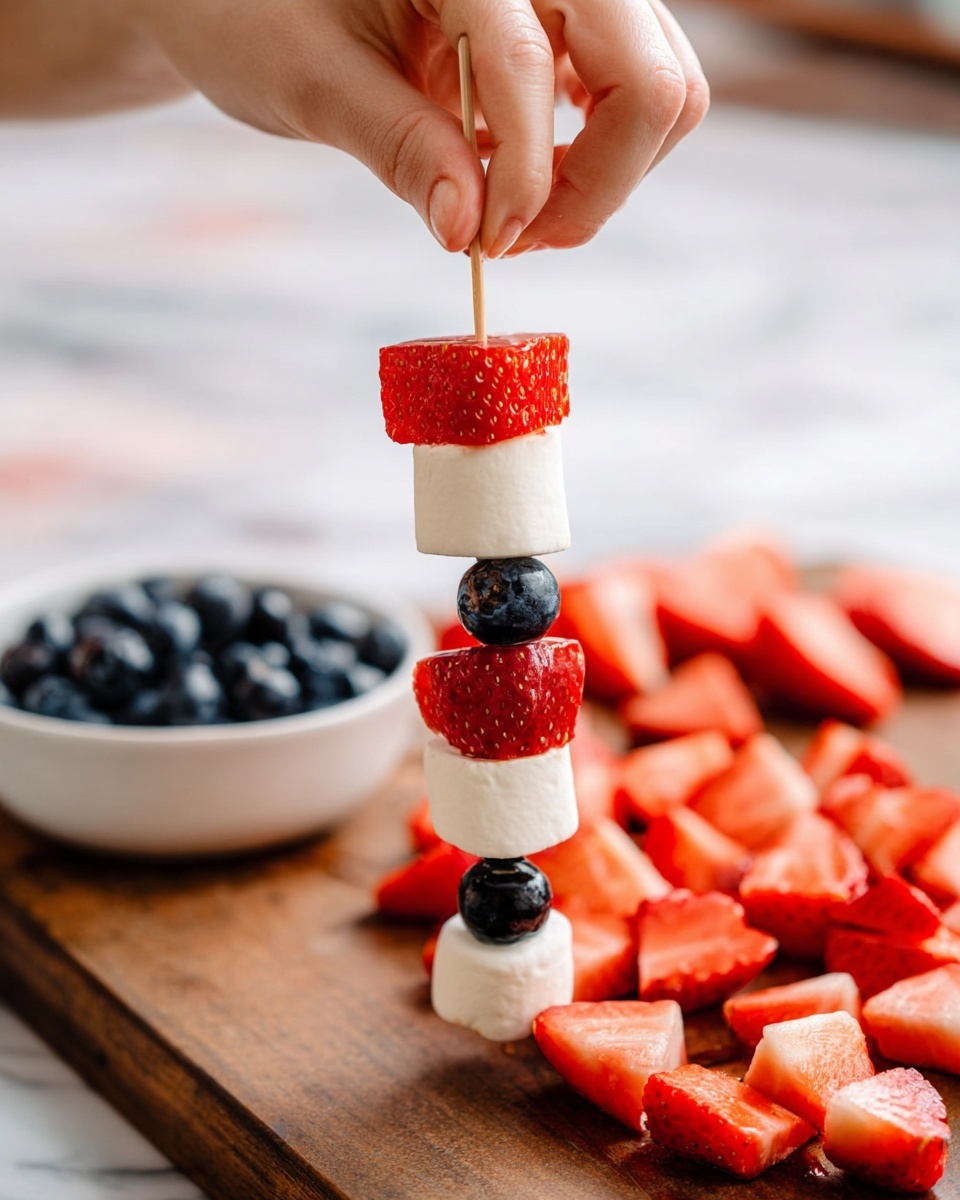 A woman's hand is holding a skewer with a pattern of six layers: starting from the bottom, a red strawberry piece with seeds, then a white marshmallow square, a dark blue blueberry, a red strawberry piece, a white marshmallow square, a dark blue blueberry, and a red strawberry piece at the top. In the background, there is a wooden board covered with cut red strawberry pieces and a white bowl filled with dark blue blueberries on a white marbled surface. The focus is on the skewer and the woman's hand placing the last marshmallow piece. Photo taken with an iphone --ar 4:5 --v 7