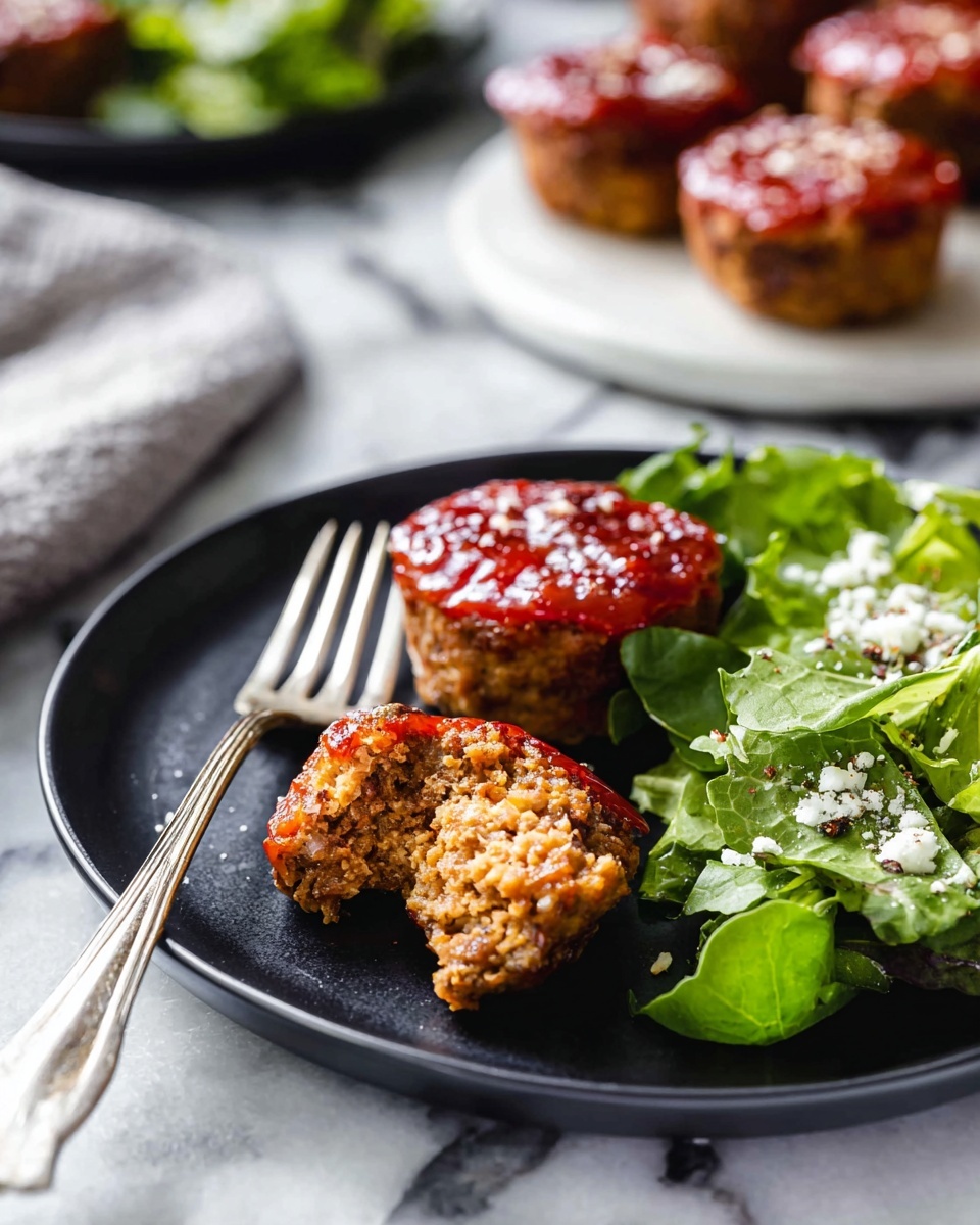 The image shows a close-up of three small round meatloaf muffins on a dark plate, each topped with a shiny reddish glaze. One muffin is broken in half, revealing a brown, tender, and crumbly inside, with a small piece on a silver fork to the left. On the right side of the plate, there is a small pile of green leafy salad sprinkled with white cheese crumbles and a little black pepper. In the blurred background, there is a white marbled surface and another plate with more meatloaf muffins. Photo taken with an iphone --ar 4:5 --v 7