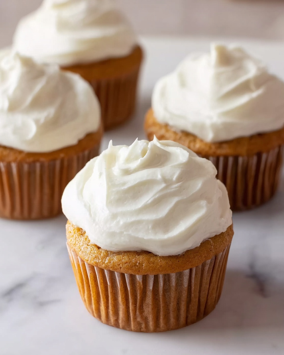 The image shows four cupcakes placed on a white marbled surface. Each cupcake has one visible cake layer in a light golden brown color with a smooth but lightly textured surface. On top of each cake layer is a thick layer of white frosting, spread evenly with soft, swirling patterns that create a fluffy and creamy look. The cupcakes are in plain, brown paper liners. The focus is on the cupcake in the front center, with the other three slightly blurred in the background. photo taken with an iphone --ar 4:5 --v 7