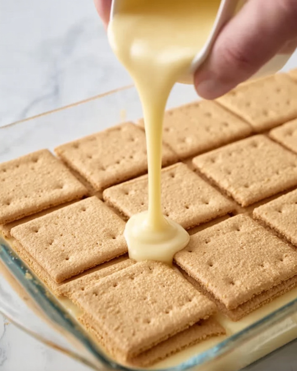 A woman's hand is pouring a smooth, light yellow creamy sauce over a single layer of square-shaped graham crackers arranged neatly in a glass dish. The graham crackers have small perforations and a light brown, crumbly texture. The sauce slightly pools on top of the crackers as it is being poured, creating a glossy finish. The background and surface are white marble, giving a clean and bright look. Photo taken with an iphone --ar 4:5 --v 7