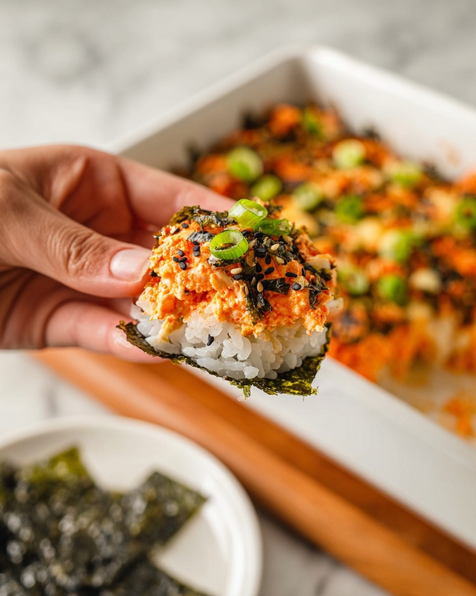 A woman's hand is holding a piece of food wrapped in dark green seaweed, with visible layers including white rice at the bottom, topped with a bright orange creamy mixture, small green onion slices, and black sesame seeds sprinkled on top. In the background, there is a white rectangular baking dish containing a similar layered dish with white rice as the base, the orange creamy layer with green onions and black sesame seeds spread on top. A white marbled texture surface is beneath the dish, and a white plate with more dark green seaweed pieces is blurred in the background. photo taken with an iphone --ar 4:5 --v 7