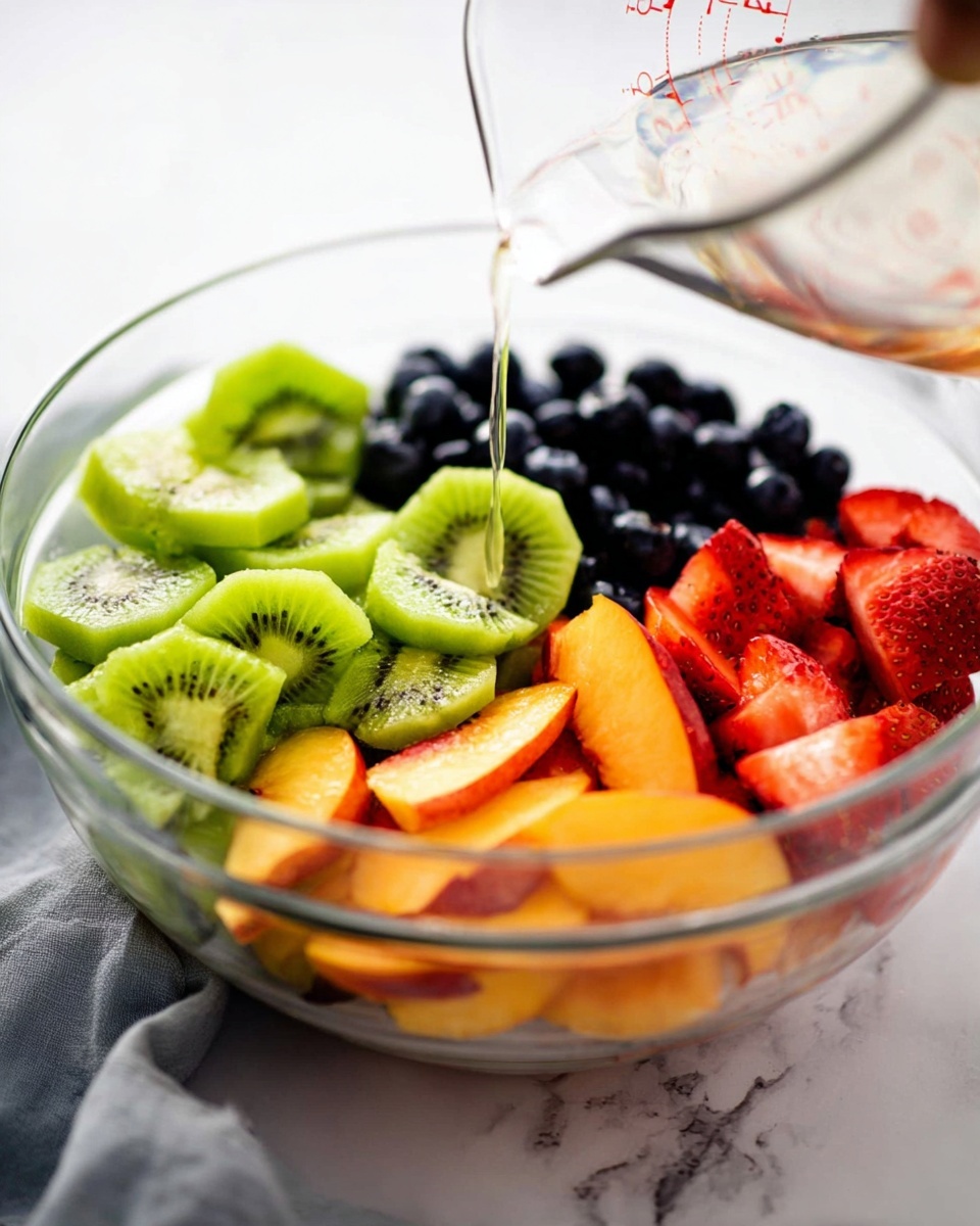 A clear glass bowl filled with four main fruit layers arranged in sections: bright green sliced kiwi with small black seeds on the left, dark round blueberries grouped at the top, red strawberry slices with seeds showing at the bottom left, and orange peach slices with smooth skin on the right. A transparent liquid is being poured over the kiwi from a clear measuring cup held just above the bowl. The bowl is set on a white marbled surface with a soft gray cloth blurred in the background. Photo taken with an iphone --ar 4:5 --v 7