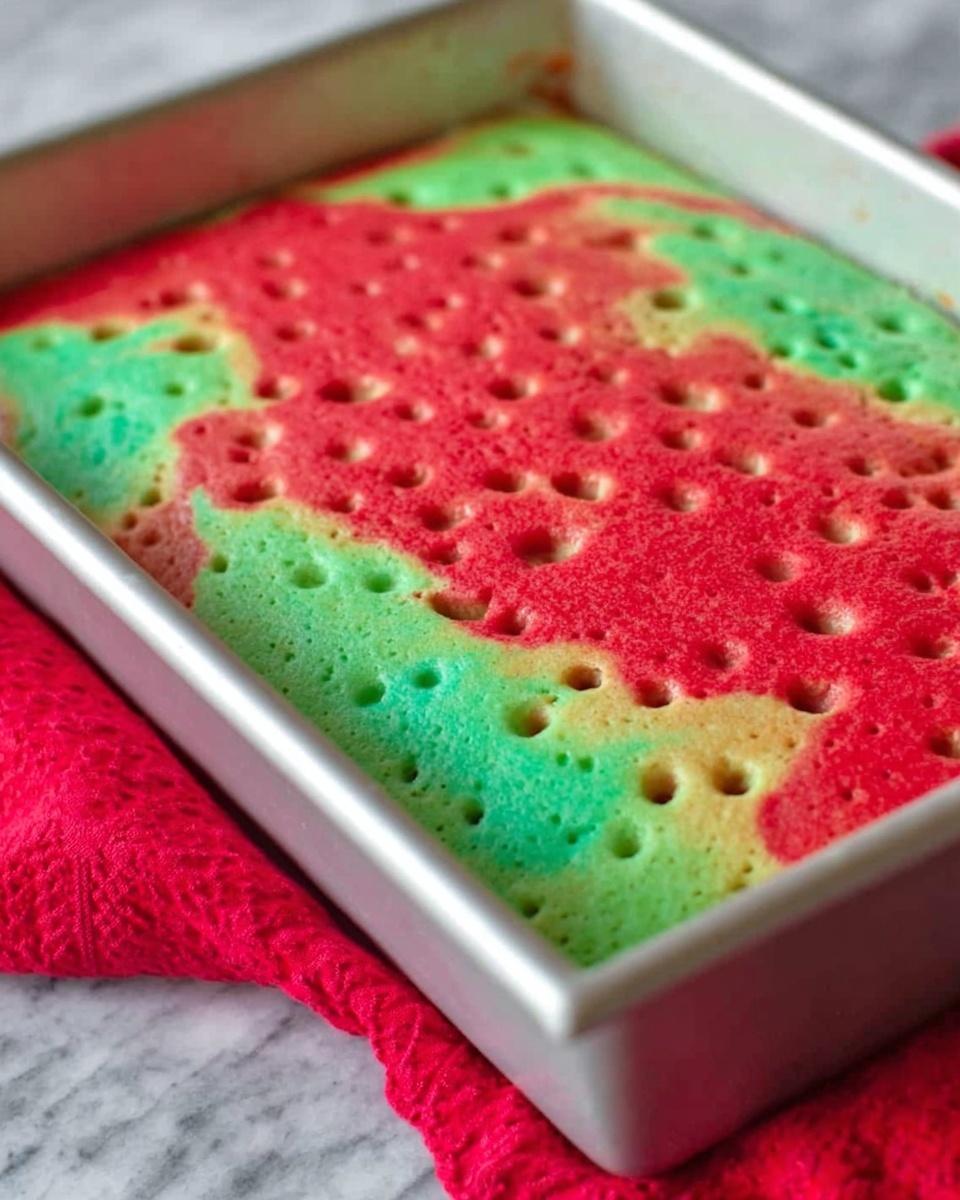 A close-up image of a rectangular cake in a white baking pan showing two main colorful layers. The top layer is a marbled mix of bright red and green with small holes poked evenly across the surface. The cake has a light, soft texture with some spots where darker bits peek through under the colors. The pan sits on a white marbled surface partially covered with a red cloth underneath. Photo taken with an iphone --ar 4:5 --v 7
