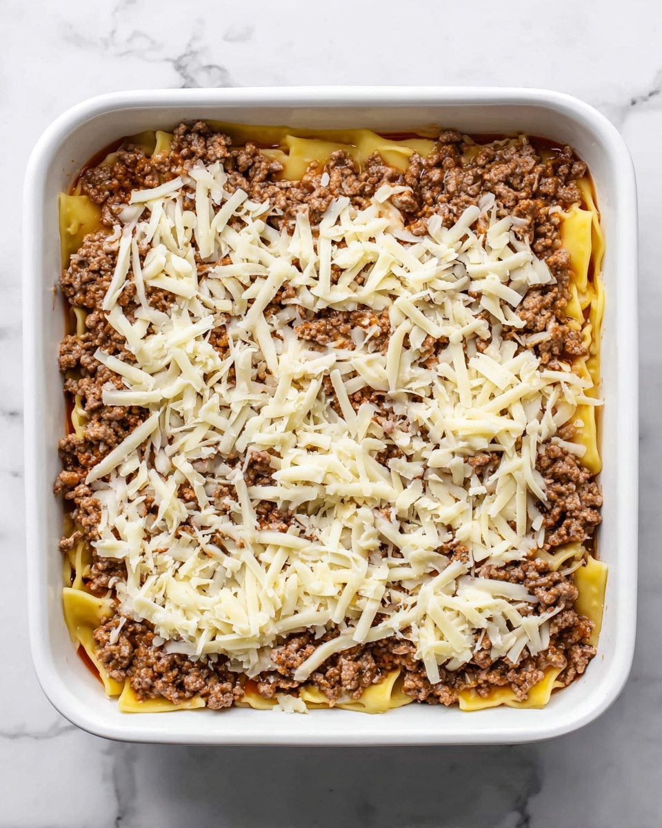 The image shows a square white baking dish on a white marbled surface filled with a layered dish. The bottom layer consists of yellow pieces that look like cooked pasta or slices of a yellow vegetable. On top of this is a layer of brown cooked ground meat spread evenly across the dish. The final layer consists of scattered white shredded cheese spread over the meat. The cheese strands are uneven and loosely placed, giving a rough texture to the top. photo taken with an iphone --ar 4:5 --v 7