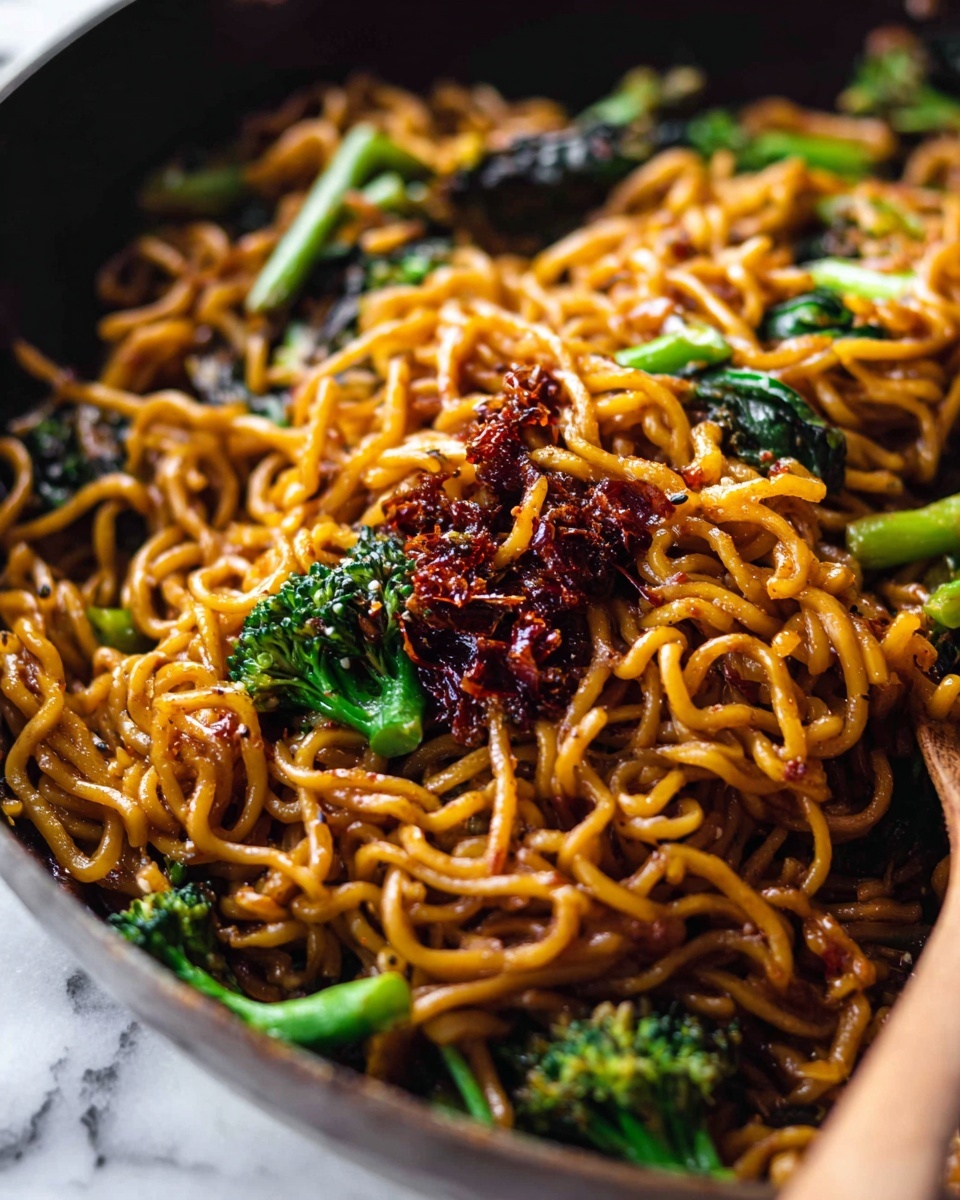 The image shows a close-up of a pan filled with cooked curly noodles coated in a shiny, brown sauce. Mixed in are bright green broccoli florets and some dark leafy greens, adding contrast and texture. On top of the noodles, there are chunky pieces of a reddish-brown chili paste with visible seeds, adding a hint of spice and rich detail. The noodles look soft and twisted together, while the chili paste and vegetables provide rough, uneven textures. The background is a white marbled surface, and a wooden spoon is partially visible on the right side of the pan. photo taken with an iphone --ar 4:5 --v 7