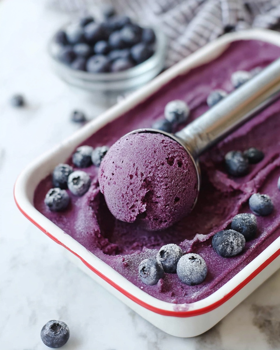 A white rectangular dish with a red rim is filled with a smooth, deep purple frozen dessert. On the surface, whole blueberries are scattered, some frosted, adding dark purple and blue tones. A metal ice cream scoop is lifting a round, well-shaped ball of the purple dessert from the dish, showing a creamy texture with small specks. The dish sits on a white marbled surface, and in the background, a small clear bowl filled with fresh blueberries is slightly blurred. photo taken with an iphone --ar 4:5 --v 7