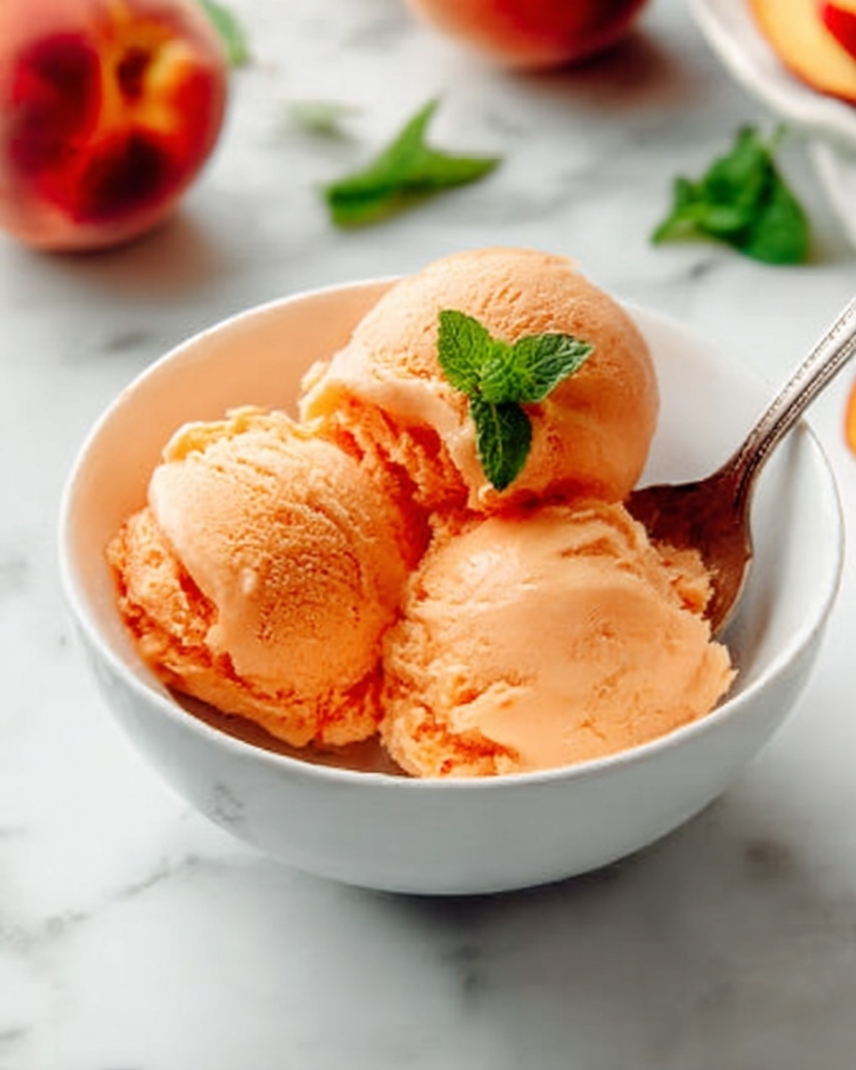 A white bowl with three smooth scoops of light orange ice cream, each scoop showing soft texture and slight melting edges. A small green mint leaf rests on top of the front scoop. A silver spoon is placed inside the bowl, with a woman's hand holding the spoon's handle. The bowl sits on a white marbled surface, with blurred peaches and green leaves in the background. photo taken with an iphone --ar 4:5 --v 7