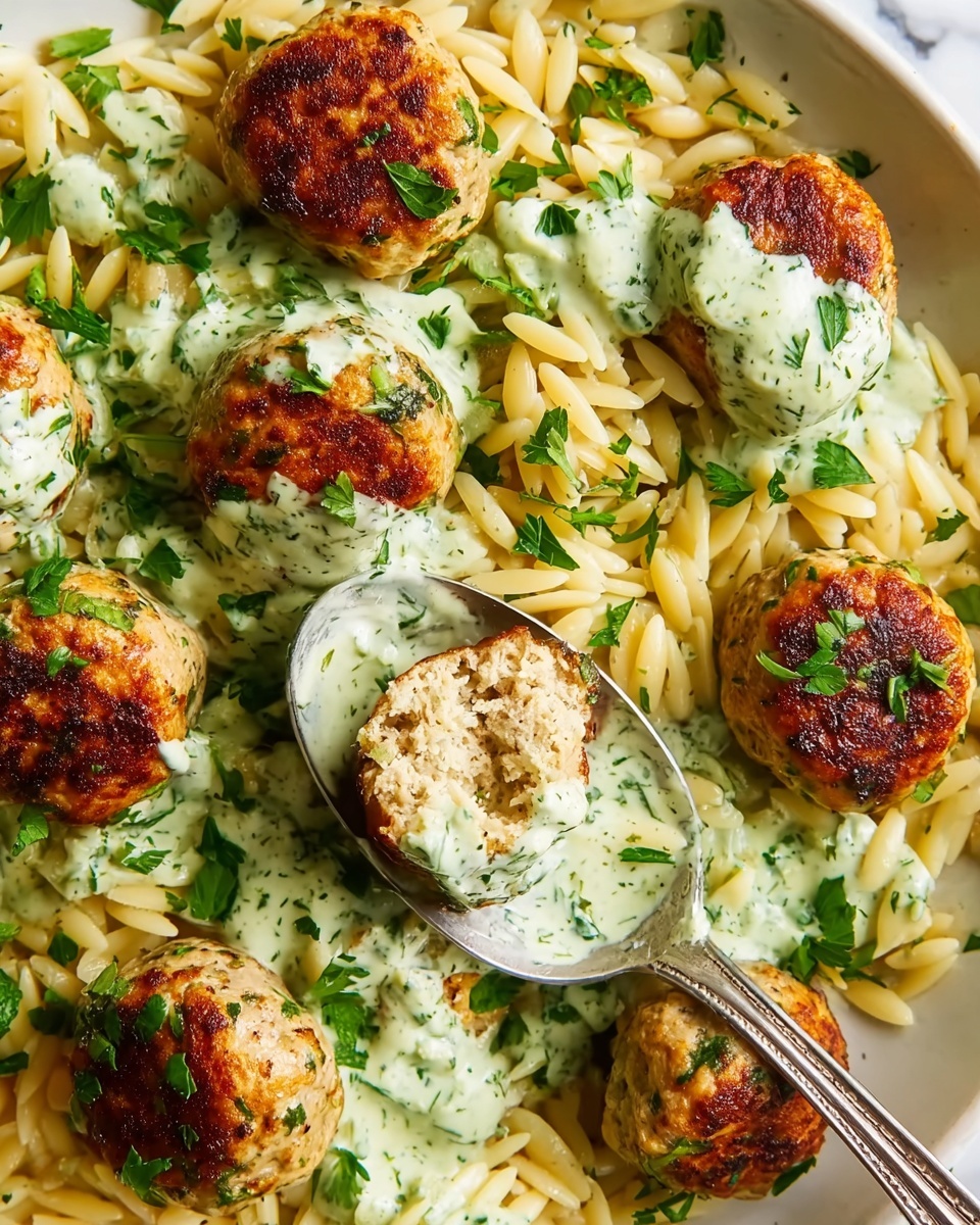 The image shows a close-up of a white plate filled with light golden orzo pasta and round, golden-brown meatballs. There are eight meatballs in total, with one cut in half revealing a light, moist inside. The meatballs are topped with a creamy, pale green herb sauce that has small green flecks. Fresh green parsley leaves are scattered on top and between the layers of orzo and meatballs. A silver spoon rests on the plate, holding orzo and a sauce-covered meatball half. The background is a white marbled texture. Photo taken with an iphone --ar 4:5 --v 7