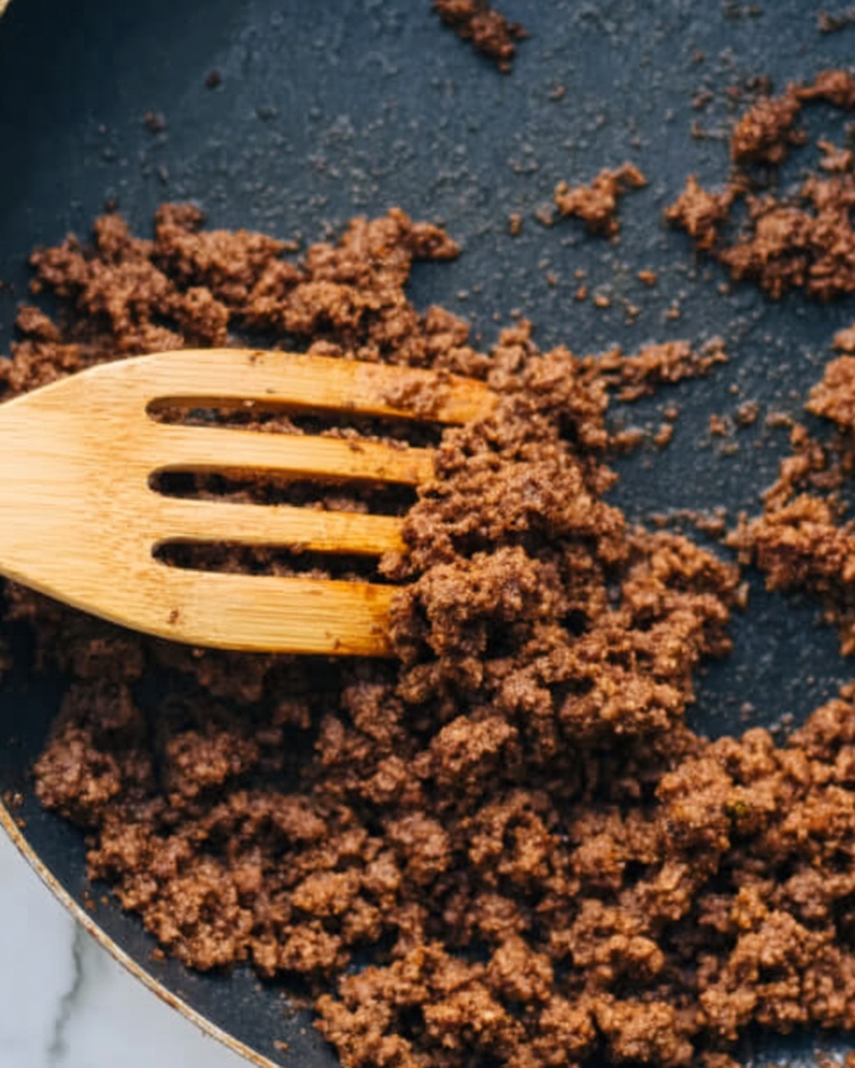A close-up view of a frying pan filled with cooked ground meat mixed with small chopped green peppers and onions. The mixture has a brown and slightly glossy texture, showing cooked, crumbly ground meat with soft, slightly translucent vegetable pieces evenly spread throughout. A woman's hand is holding a wooden spoon stirring the food inside the pan. The pan sits on a black stove with a white marbled surface visible around. Photo taken with an iphone --ar 4:5 --v 7