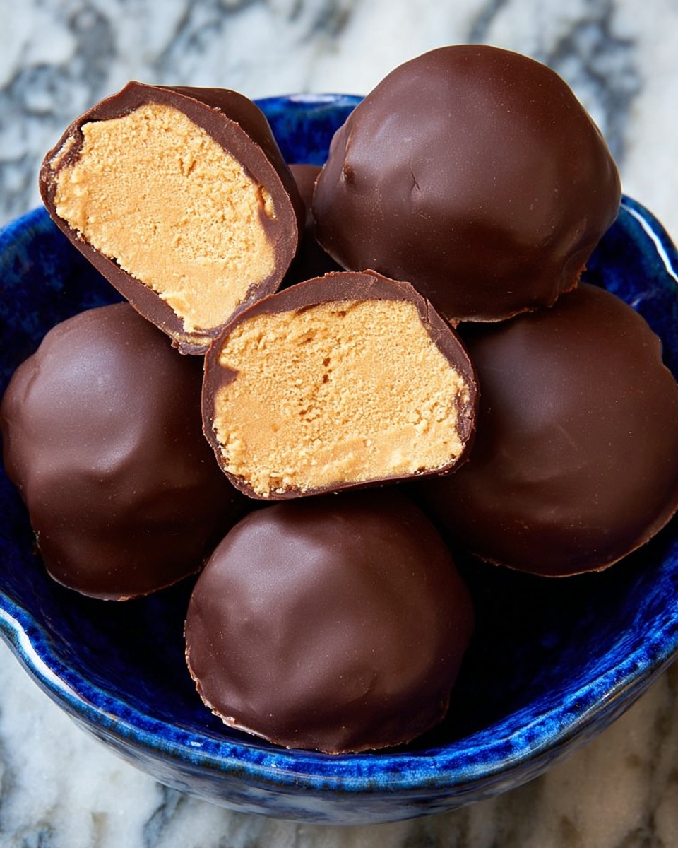 A blue patterned bowl filled with round chocolate-covered spheres, three whole and two sliced to show their inside. Each sphere has two layers: a smooth, dark brown outer chocolate shell and a light tan, slightly crumbly-looking interior filling. The spheres sit close together, filling the bowl, which has a shiny finish. The background is a white marbled texture. Photo taken with an iphone --ar 4:5 --v 7
